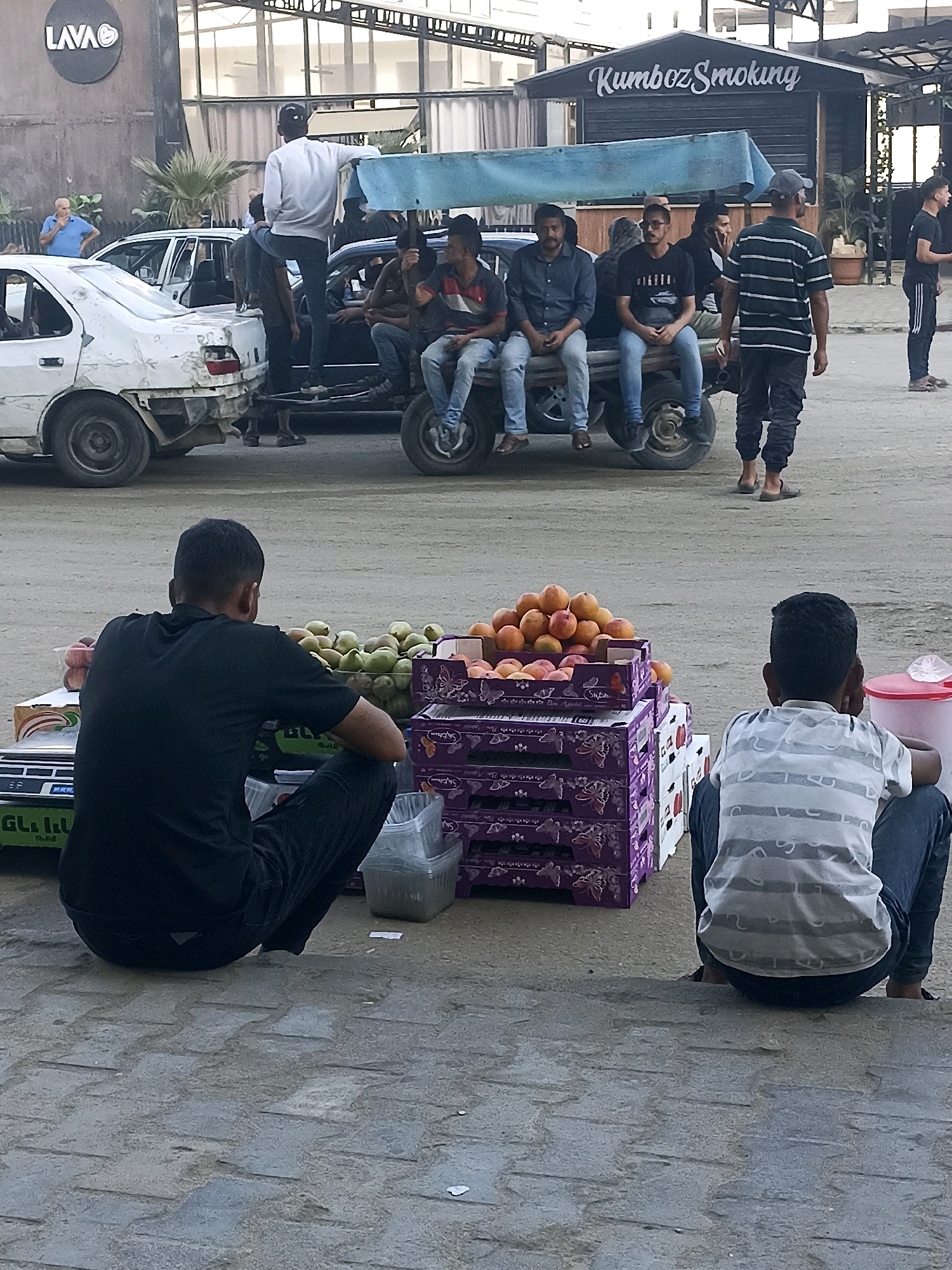 In the city of Gaza, Palestinian Territories, a street scene unfolds with several individuals and vehicles. In the background, a building with "LAVA" signage and another with "Kumbol Smoking" lettering are visible, suggesting a commercial area. A white car with visible damage, possibly a Peugeot, is parked on the left. A dark-colored car is also present, carrying several men. One man, wearing a white long-sleeved shirt and dark pants, stands on the back of the dark car, facing away from the camera. Two other men are seated on the back of the dark car, one in a blue shirt and the other in a dark shirt. A man in a striped shirt and dark pants walks in the middle ground, and another man in a black shirt and dark pants stands to his right. In the foreground, two boys are seated on the ground. The boy on the left is wearing a black shirt and dark pants, facing a fruit stand. The boy on the right is wearing a striped t-shirt and dark pants, facing away from the camera. Various fruits, including peaches and what appear to be pears or apples, are displayed in boxes on the fruit stand. A red cooler is also near the fruit stand. The ground is paved with grey cobblestones.