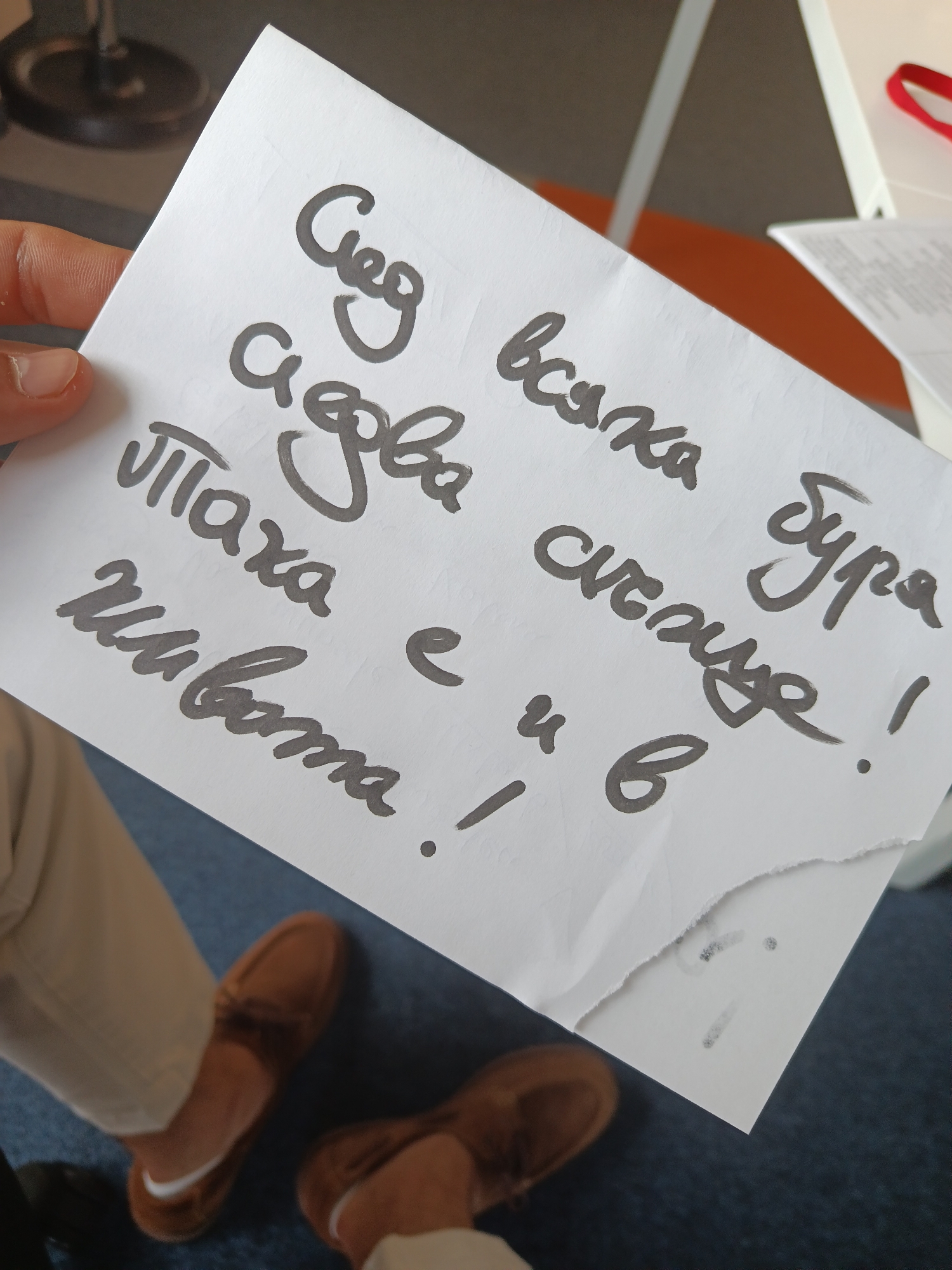 A close-up, slightly angled shot captures a person's hand holding a white piece of paper with handwritten text. The paper is somewhat creased, and its bottom-right corner is torn.
In the background, out of focus, are the lower legs of the person, clad in light-colored (beige or khaki) pants, and their feet, wearing brown loafer-style shoes. A white detail, possibly part of a sock or shoe lining, is visible near the ankle of the left foot. The floor beneath is covered with a blue carpet. Other blurry elements in the background suggest an indoor setting, possibly an office or event space, with some white furniture and a red strap or ribbon visible.
The prominent feature is the handwritten text on the white paper, written in dark gray or black ink with a flowing, informal script. The text is in Bulgarian and reads:
"След всяка буря
сядва
Паха е в
живота !"
Translated literally, this means:
"After every storm
sits (down/settles)
Paha is in
life!"
This message appears to be a personal note, possibly an encouraging sentiment about resilience after difficulties, potentially addressed to someone named "Paha." The scene takes place indoors in Varna, Bulgaria. No specific time of day, weather, or explicit emotions are discernible from the image.