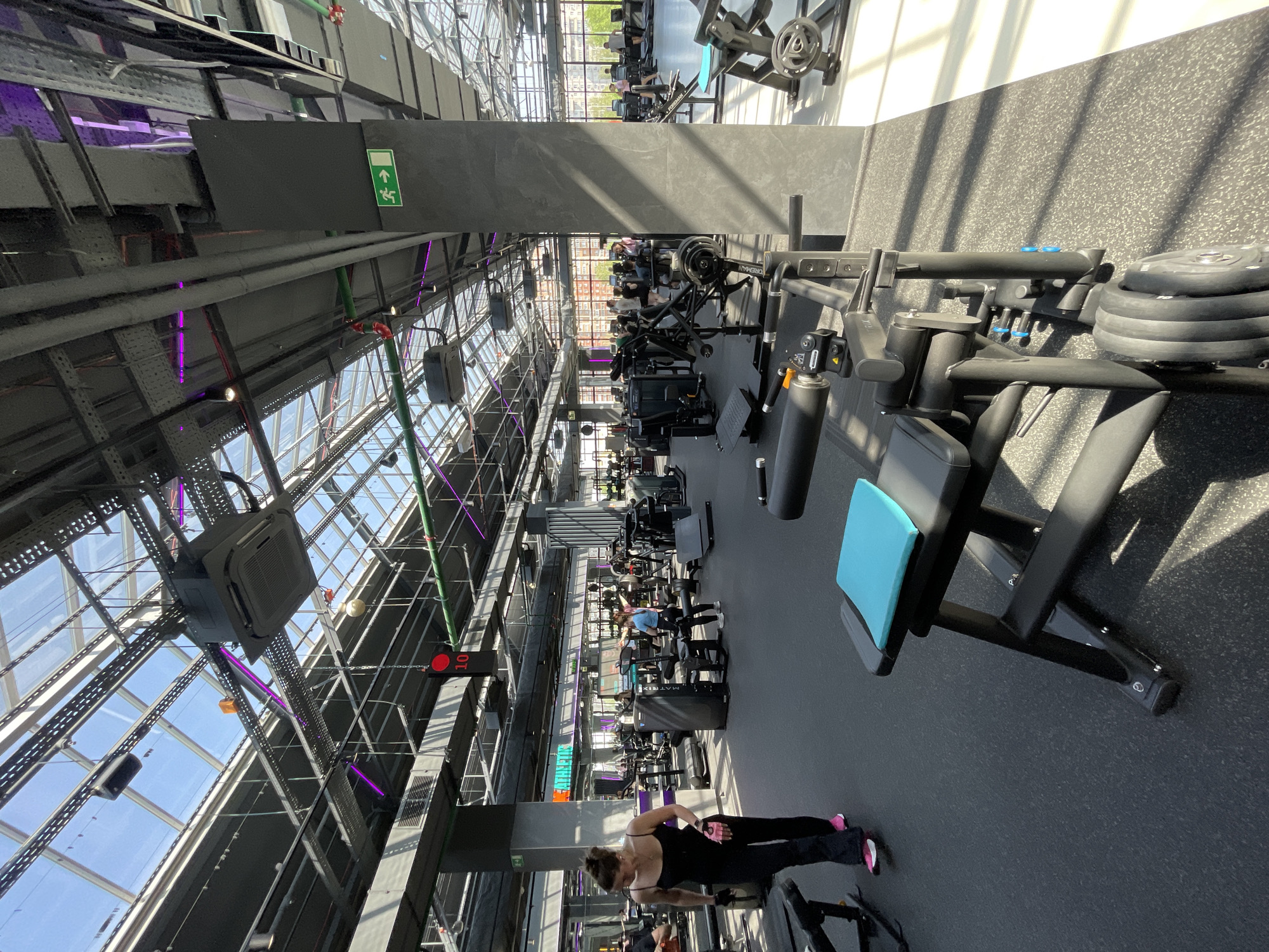 The image depicts the interior of a well-lit gymnasium located in Kazan, Russia. The gym is equipped with a variety of exercise machines, including weightlifting stations and leg press machines. The building's structure features an exposed ceiling with metal beams, ductwork, and lighting fixtures, with large windows allowing natural light to enter. Gym patrons are seen exercising. One woman in the foreground is performing a weightlifting exercise. The overall scene captures a typical modern gym environment.