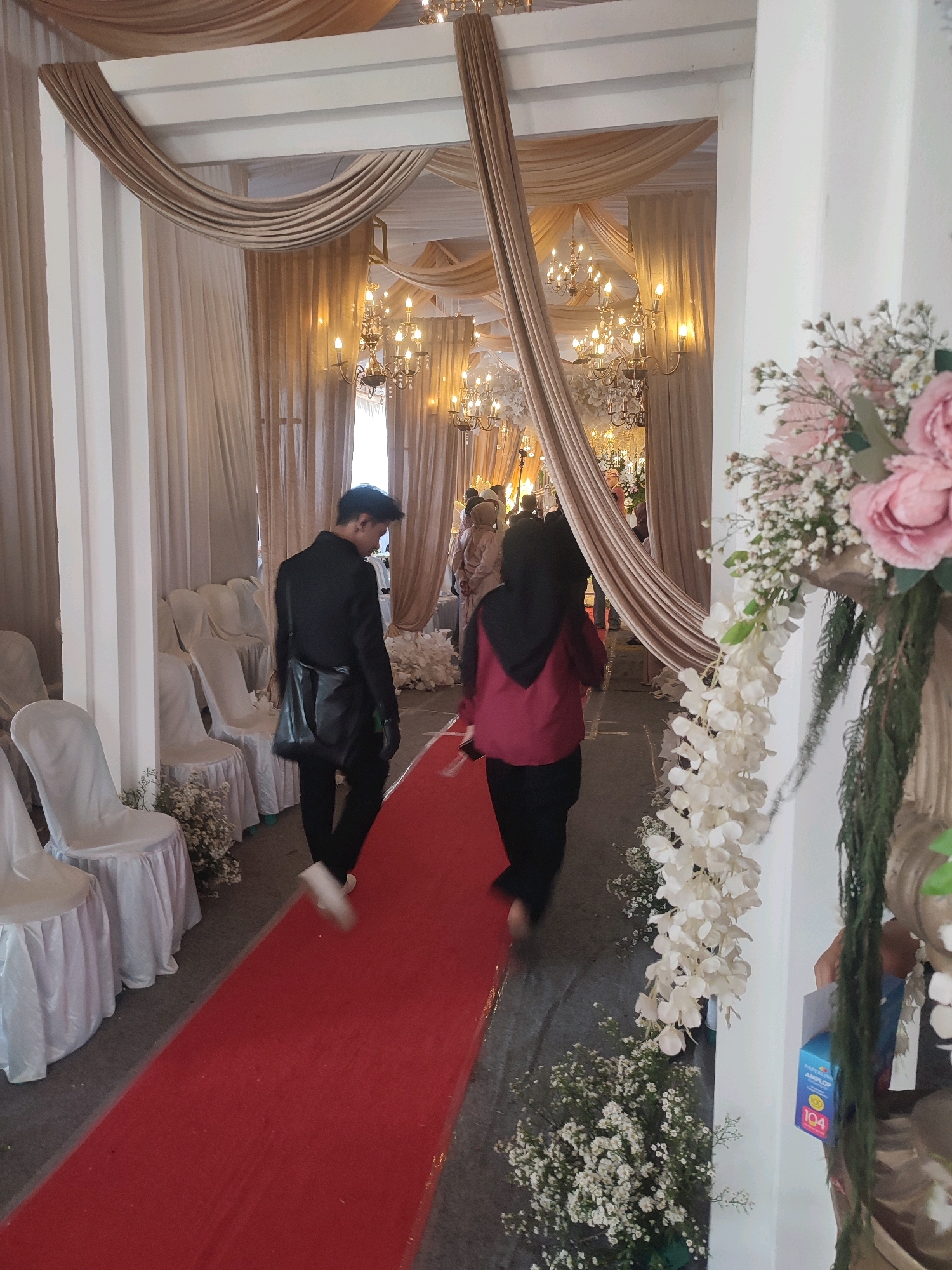 The image depicts an indoor wedding ceremony setup in what appears to be a hall or reception venue in Kecamatan Haurgeulis, Indonesia. The scene is decorated with draped beige and tan fabric on the ceiling, creating a canopy effect, and numerous chandeliers. White chairs, covered with fabric, are arranged in rows on the left side of a prominent red carpet runner. Floral arrangements of white and pink flowers are visible throughout the venue, with a large arrangement featuring pink roses and white cascading flowers on the right side of the image.
In the foreground, two individuals are walking along the red carpet. The person on the left is a man wearing a black suit and a black messenger bag, walking away from the camera. The person on the right is a woman wearing a maroon jacket with a black headscarf and black pants, also walking away from the camera. In the background, other guests are visible, some wearing headscarves, suggesting a Muslim wedding ceremony. The lighting appears to be warm and ambient, provided by the chandeliers. A blue box with text "AMPLOP 104" is visible on the right. The floor is a dark gray surface around the red carpet.