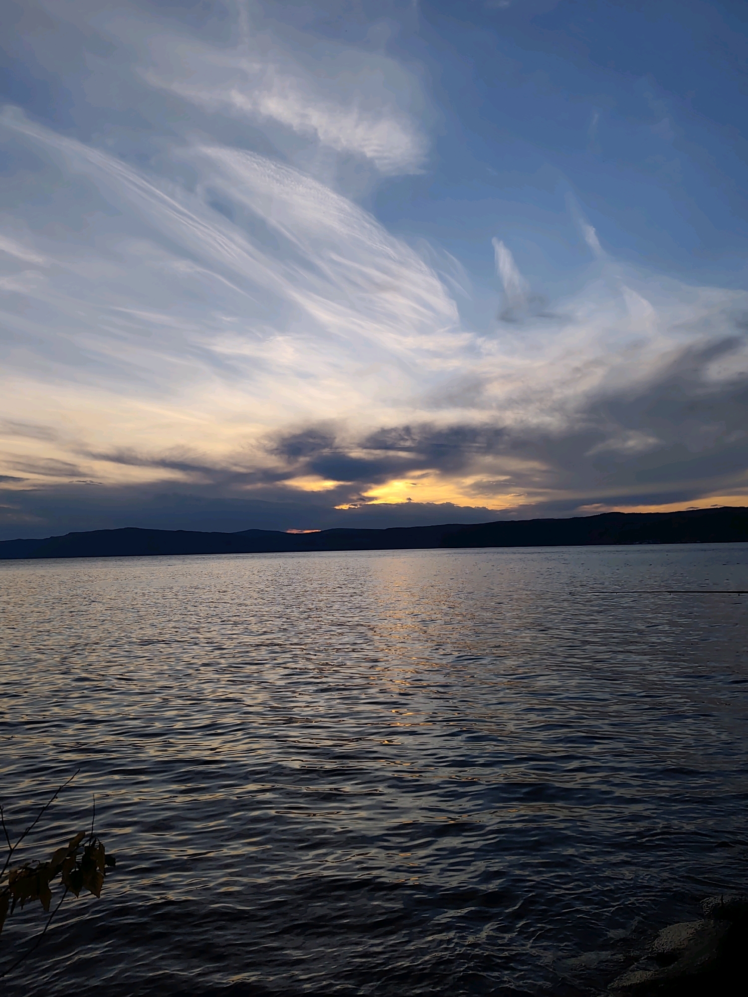 A serene lake reflects the dusky sky as the sun sets behind a silhouetted mountain range. The water's surface is rippled, with subtle glints of light catching the waves. Wispy clouds stretch across the vast expanse of the sky, painted with hues of blue, orange, and gray. In the foreground, a few bare branches with wilting leaves hang near the water's edge, and a hint of rocky shore is visible on the right. The scene evokes a sense of tranquility and the fading light of day.