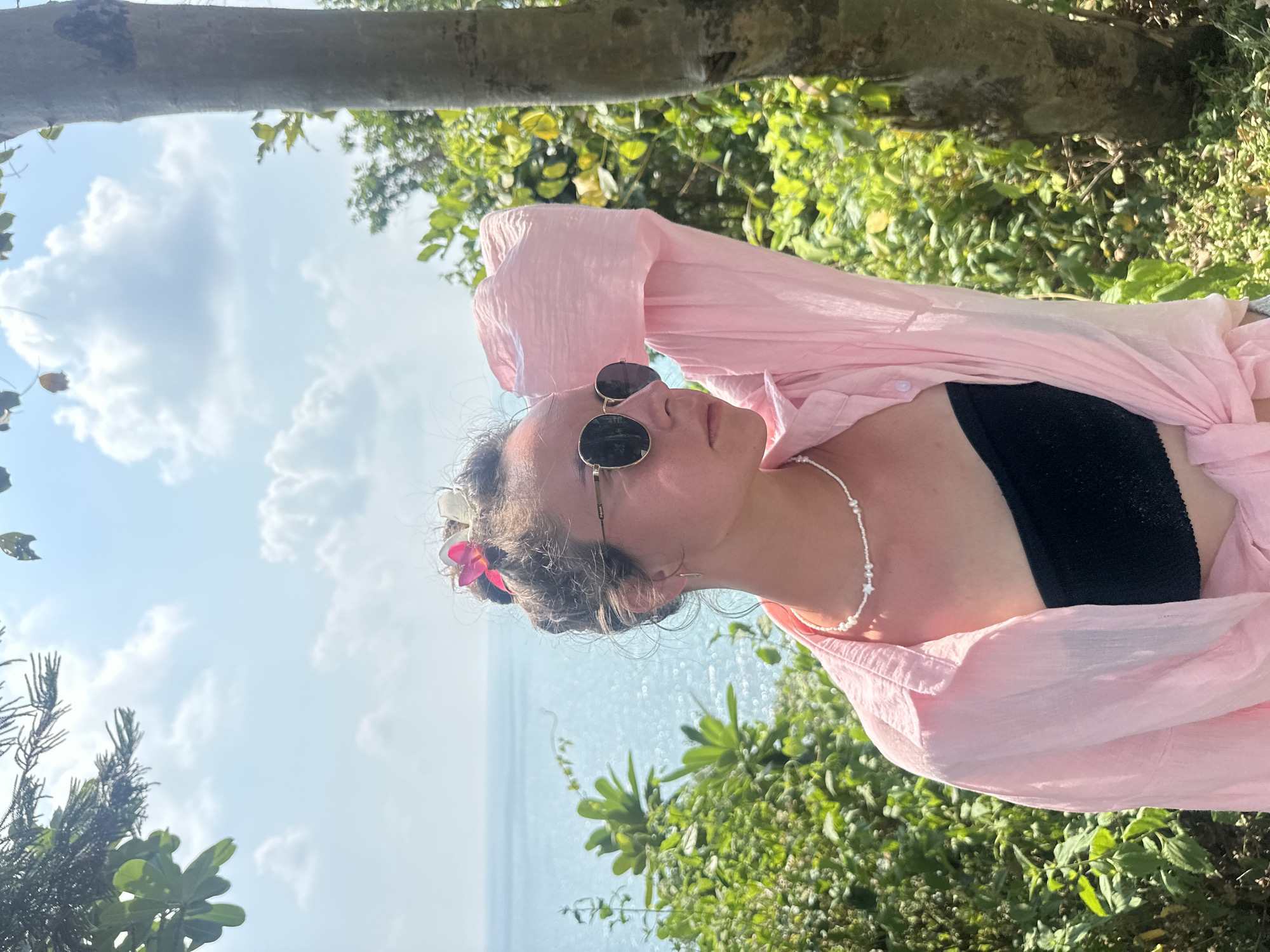 A woman is lying in an outdoor setting in Ao Hin Khok, Thailand. She is wearing a black bikini top and a light pink, unbuttoned shirt. The woman is also wearing sunglasses and a pearl necklace. There is a pink flower in her hair. The background shows a blue sky with scattered clouds and the ocean, along with lush green vegetation. A tree branch is positioned across the top of the frame.