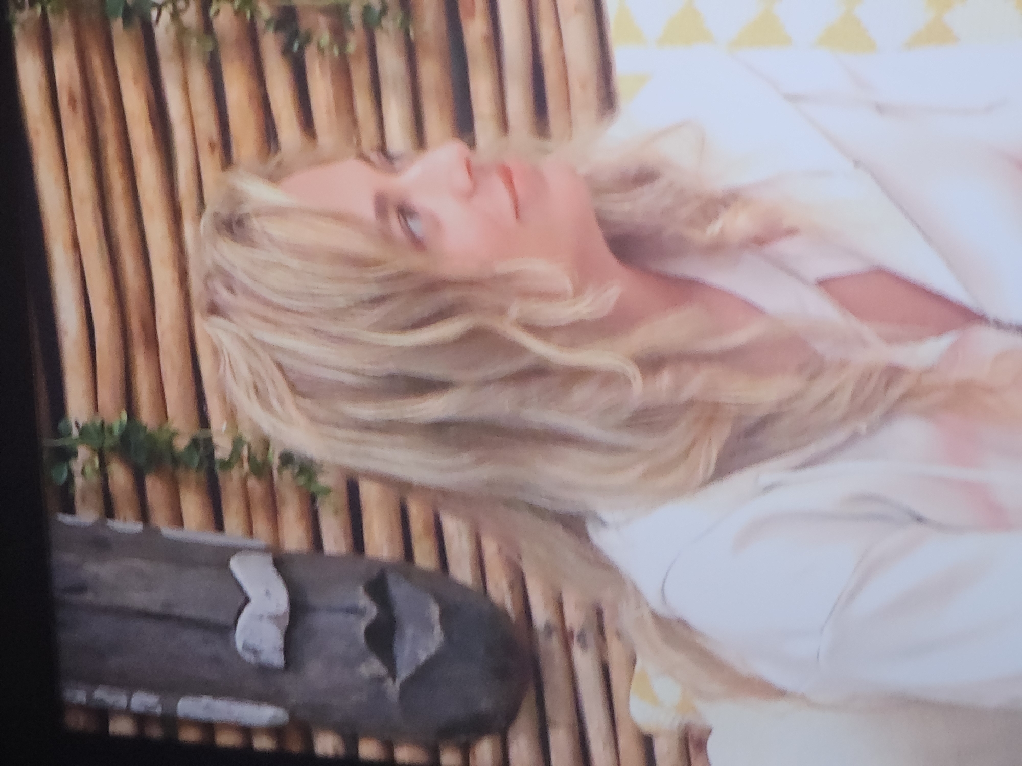 The image shows a medium shot of a woman with blonde, wavy hair, wearing a white zip-up hoodie. She is looking to her right with a slight smile. The background features a wall constructed of horizontal wooden planks, with a wooden tiki-style mask on the left side and a vine with green leaves hanging down from the top. A portion of a yellow and white patterned cushion is visible on the right. The scene appears to be indoors, possibly a living room or a lounge area, and the lighting is soft. There is no visible indication of the location being in the city of Buchen (Odenwald), Germany.