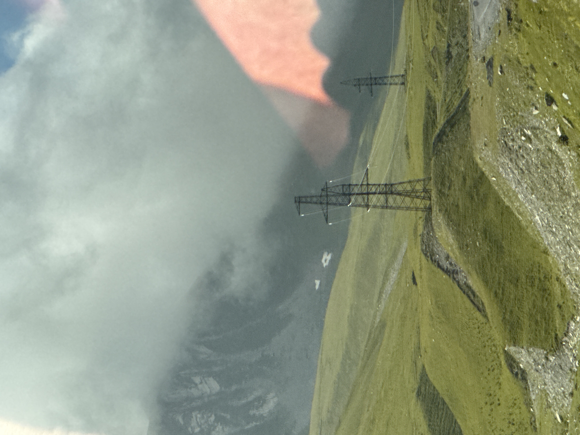 The image depicts a mountainous, grassy landscape with two electrical pylons visible. In the background, a large mountain peak is partially obscured by thick, light-colored clouds. A bright pinkish-orange light casts a shadow on the clouds and mountain. The terrain in the foreground is rolling green hills with scattered rocks. The scene appears to be set in a natural environment, possibly a rural or mountainous region, rather than a dense urban area. There are no people or specific actions depicted. The image does not provide any discernible clues or context to identify the specific location within the city of Almasiani, Georgia.