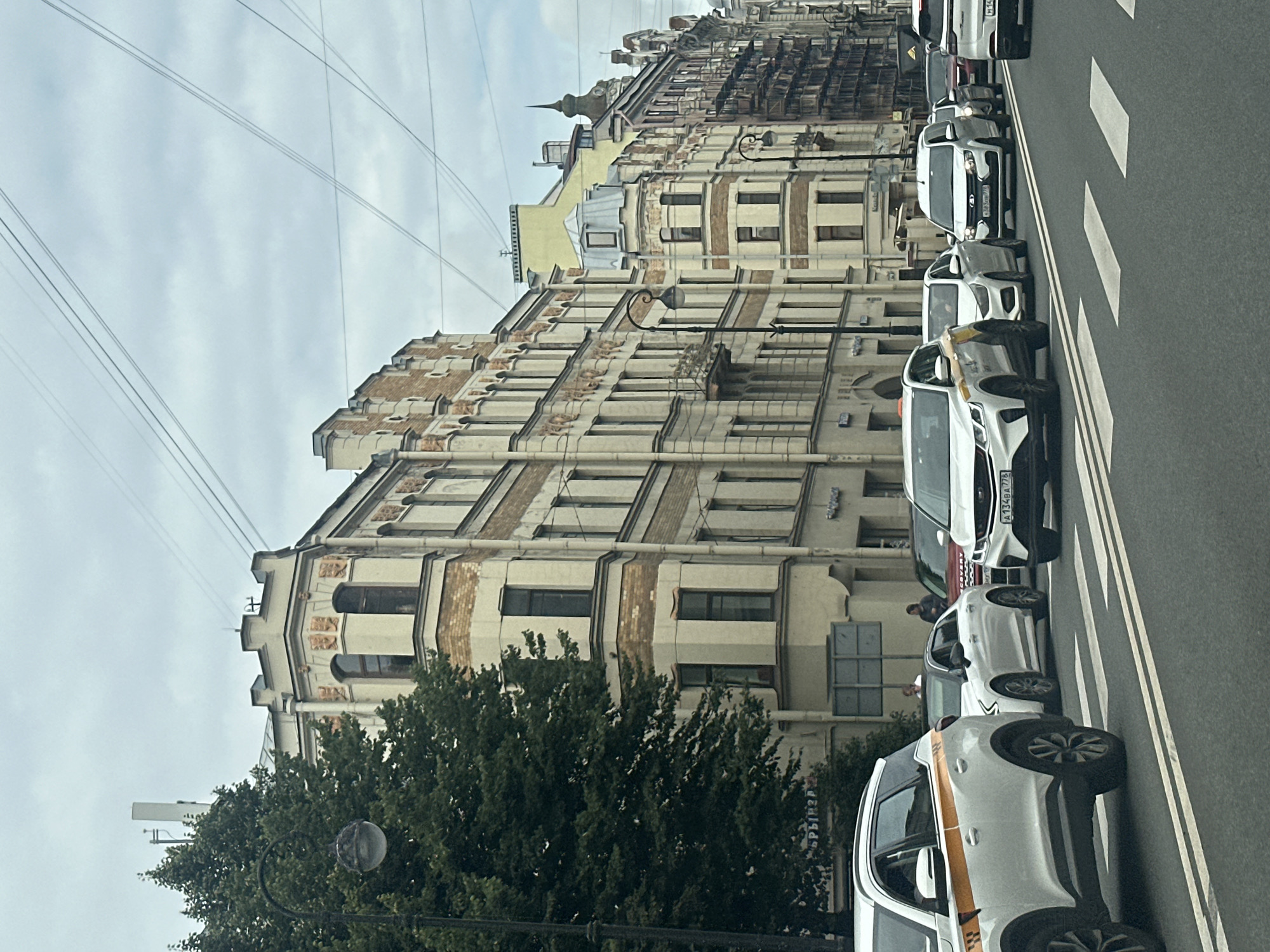 This is a medium-angle, eye-level, vertical shot of a city street in Saint Petersburg, Russia, on an overcast day. The perspective is from the perspective of a passenger in a vehicle moving through traffic. The street is paved and has double white lines marking lanes. A pedestrian crossing is visible in the foreground. A line of vehicles, including several white cars and SUVs, are stopped in traffic. The buildings lining the street are multi-story, with ornate architectural details, including decorative cornices, arched windows, and light-colored facades with some brick accents. A large, mature tree with dark green foliage is on the left side of the frame, partially obscuring a vintage-style lamppost. Overhead, a network of power lines crisscrosses the sky. The overall atmosphere is one of urban congestion under a cloudy sky.