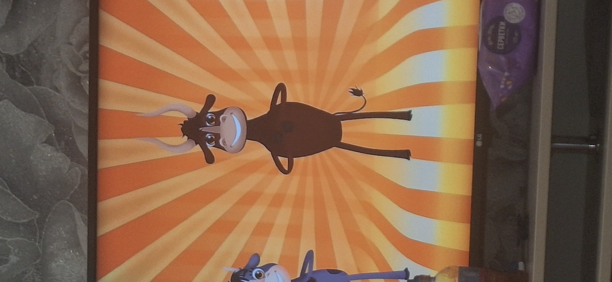 This is a photograph of a television screen displaying a cartoon image of two bulls against an orange and yellow striped background. The bull on the right is larger and brown, with large horns and a wide smile. The bull on the left is smaller and purple, with a similar expression. The TV is on a shelf, with a bottle of liquid on the left and a package of wet wipes on the right. The background behind the TV appears to be a textured wallpaper. The image does not provide any information about its location in Bila Tserkva, Ukraine.