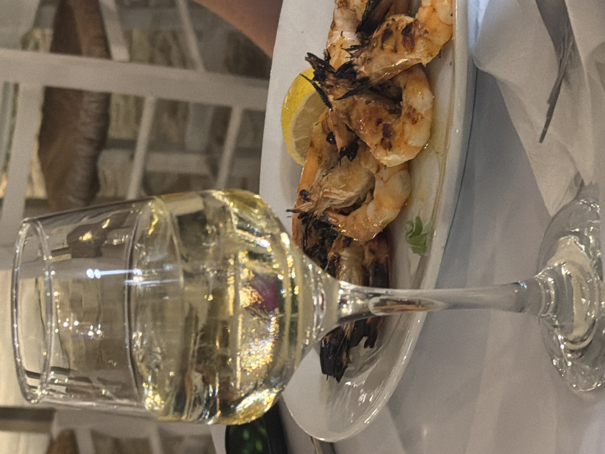 A close-up shot captures a glass of white wine, filled to about two-thirds capacity, placed in the foreground on a white tablecloth. The wine exhibits a pale yellow hue and is illuminated by a light source, creating reflections within the glass. Behind the wine glass, a white plate holds a portion of grilled shrimp and a slice of lemon. The shrimp appear to be seasoned and have grill marks. The background is slightly blurred but suggests an outdoor dining setting, possibly at a restaurant in the Sithonia Municipal Unit, Greece, with white chairs and woven seating visible. The overall scene suggests a relaxed meal, likely during daylight hours given the natural light illuminating the wine.