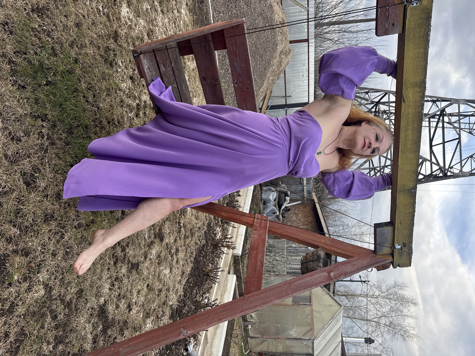 An outdoor, eye-level shot captures a woman in a vibrant purple dress against a rustic backdrop, rotated 90 degrees clockwise. The woman, with fair skin and reddish-blonde hair, is looking directly at the camera with a slight smile. She wears red lipstick and a green pendant on a dark cord around her neck. Her long, flowing purple dress features voluminous, puffy sleeves.
She is positioned with her arms extended upwards, holding onto a thick, weathered horizontal wooden beam, possibly part of a swing set or an outdoor structure. Her body is arched slightly, with her bare left leg extended forward.
The setting appears to be a backyard or a semi-rural area in Chaikovsky, Russia. The ground beneath her is covered with patchy, mostly dry, light brown grass with some sparse green growth. In the background, there's a wooden bench or part of a structure, a large pile of earth or gravel, and several small, simple buildings, possibly sheds or greenhouses. Bare tree branches are visible against the cloudy sky, and a tall metal lattice tower, resembling a power pylon or communication tower, stands prominently on the right side of the frame. The daylight suggests it's daytime, under an overcast sky. No visible text is present in the image.