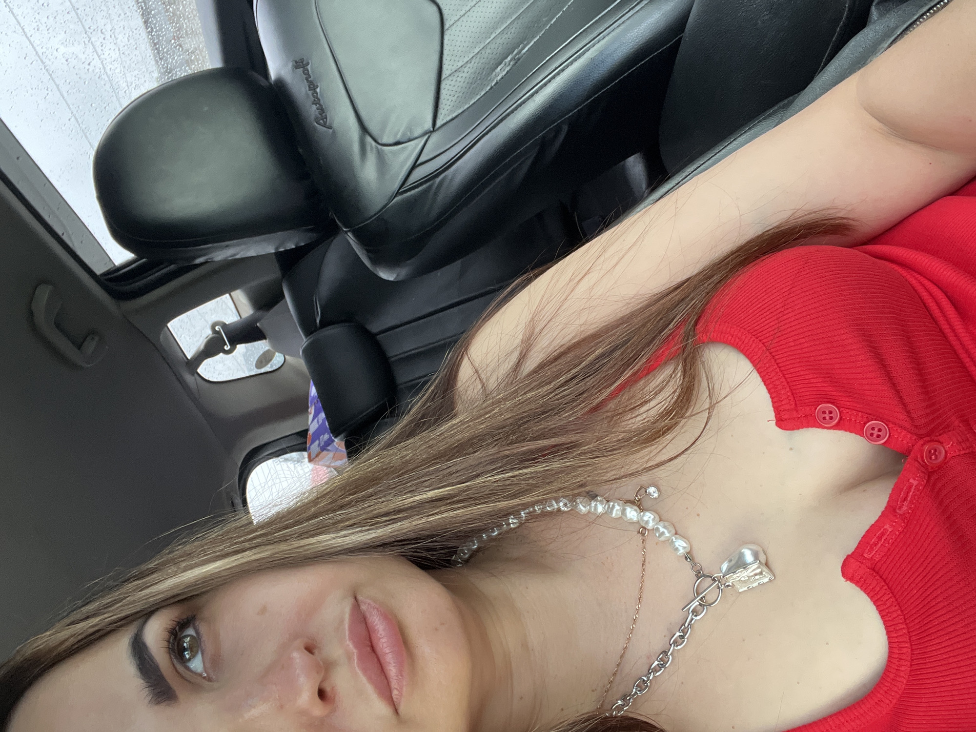 A close-up, angled selfie taken inside a car features a woman with long, light brown hair styled with highlights, which cascades over her shoulder and chest. She is wearing a red, ribbed top with several small red buttons visible along the neckline. Around her neck, she has a multi-strand pearl necklace that includes a silver toggle clasp and a textured, heart-shaped pendant, layered with a thinner, golden chain. Her face is partially visible, showing one eye looking upwards and to the left, with defined eyebrows and eyelashes. Her lips are slightly parted, indicating a relaxed expression.
The background reveals the interior of a vehicle. A black leather car seat and headrest are prominent, with the word "Autobroch" embossed on the side of the headrest. Above, a sunroof or window shows raindrops, indicating rainy or recently rainy weather outside. The overall lighting suggests it is daytime, likely overcast due to the rain. The image is taken from a low angle, emphasizing the woman's face and upper body within the car interior.
