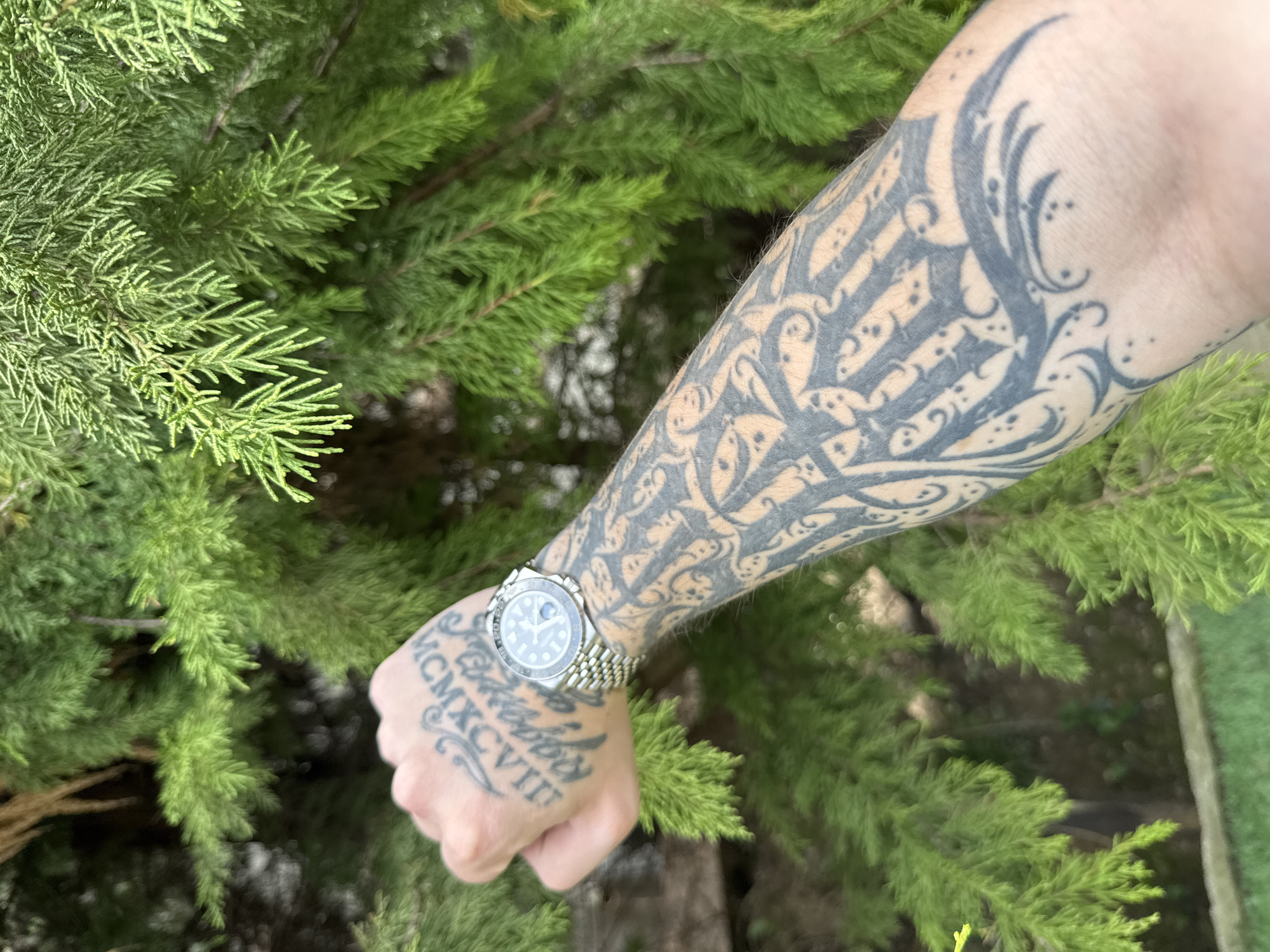 An arm with a tattoo sleeve and a watch on the wrist is positioned in front of a green coniferous tree. The tattoo extends from the wrist to the upper arm, featuring black tribal designs. The watch has a silver-colored band and a round face. The hand has a tattoo that reads "MCMXCXVII". The background consists of dense, green coniferous foliage. The location and city are unknown.