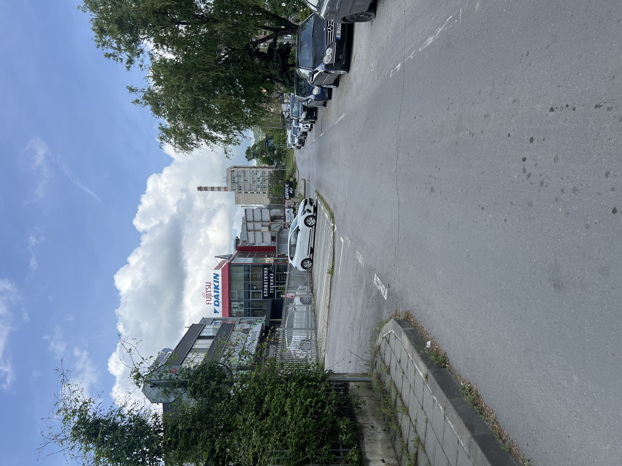 The image shows a street in Varna, Bulgaria. Buildings with "Fujitsu" and "Daikin" signage are visible on the left. A white car is parked on the side of the road next to the building. Several other cars are parked along the right side of the street, under a large tree. The road stretches into the distance, with a multi-story building visible further down the street, a chimney stands next to it. The sky is partly cloudy with a light blue color. The image is taken from a slightly elevated position. The foreground shows vegetation and a curb along the side of the road.