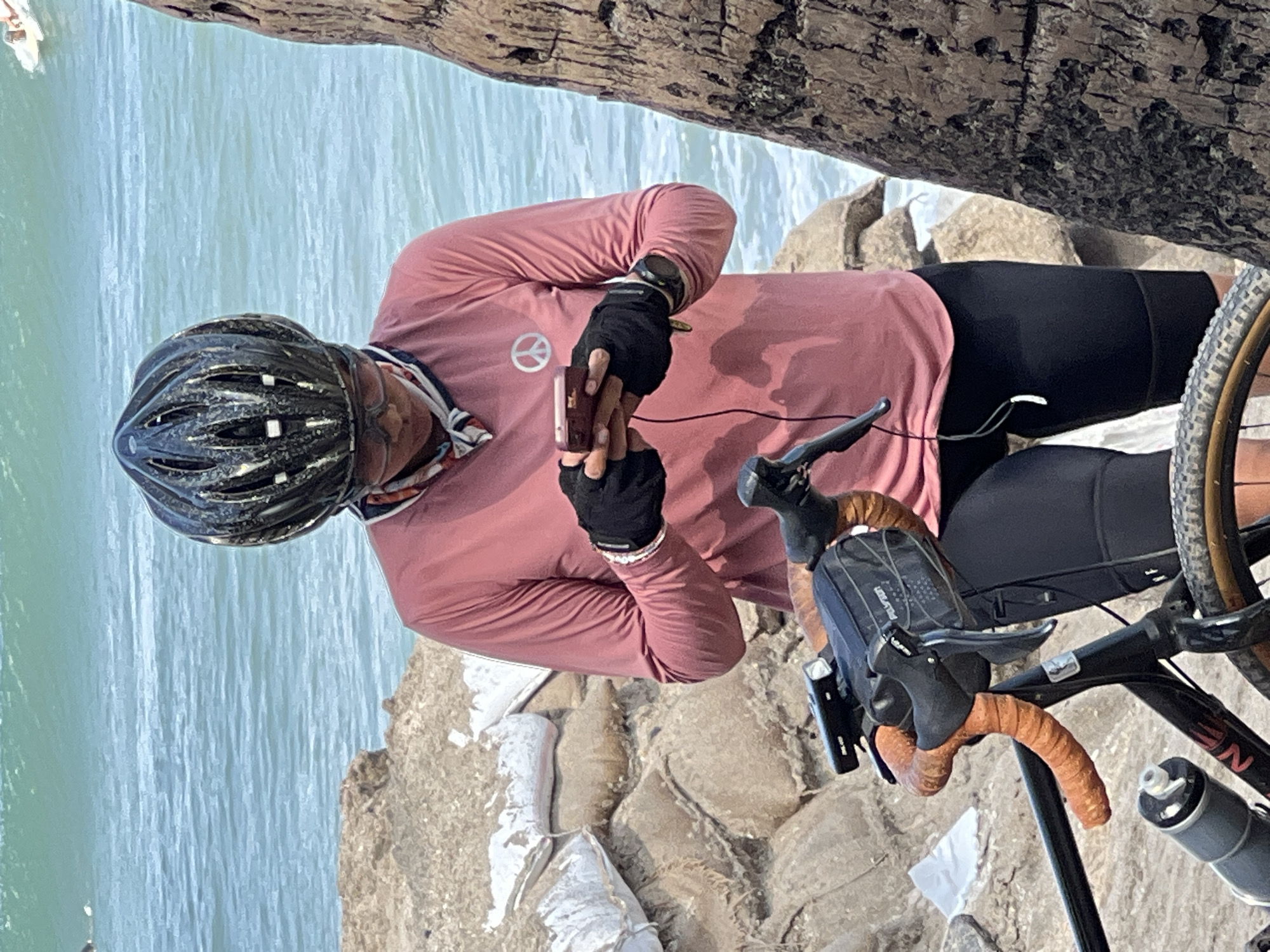 A person wearing a pink long-sleeved shirt, black cycling shorts, a black helmet, and sunglasses is standing next to a bicycle. The person is holding a smartphone and appears to be looking at it. The bicycle is black with orange handlebars and a small black bag attached to them. A water bottle is mounted on the bicycle frame. In the background, there is a body of water with waves, sandbags piled on rocks, and a portion of a tree trunk. The image is taken outdoors on a sunny day. The person's shirt has a white peace sign on it. The scene appears to be a coastal area, possibly in Kuta, Indonesia, given the presence of the ocean and palm tree.