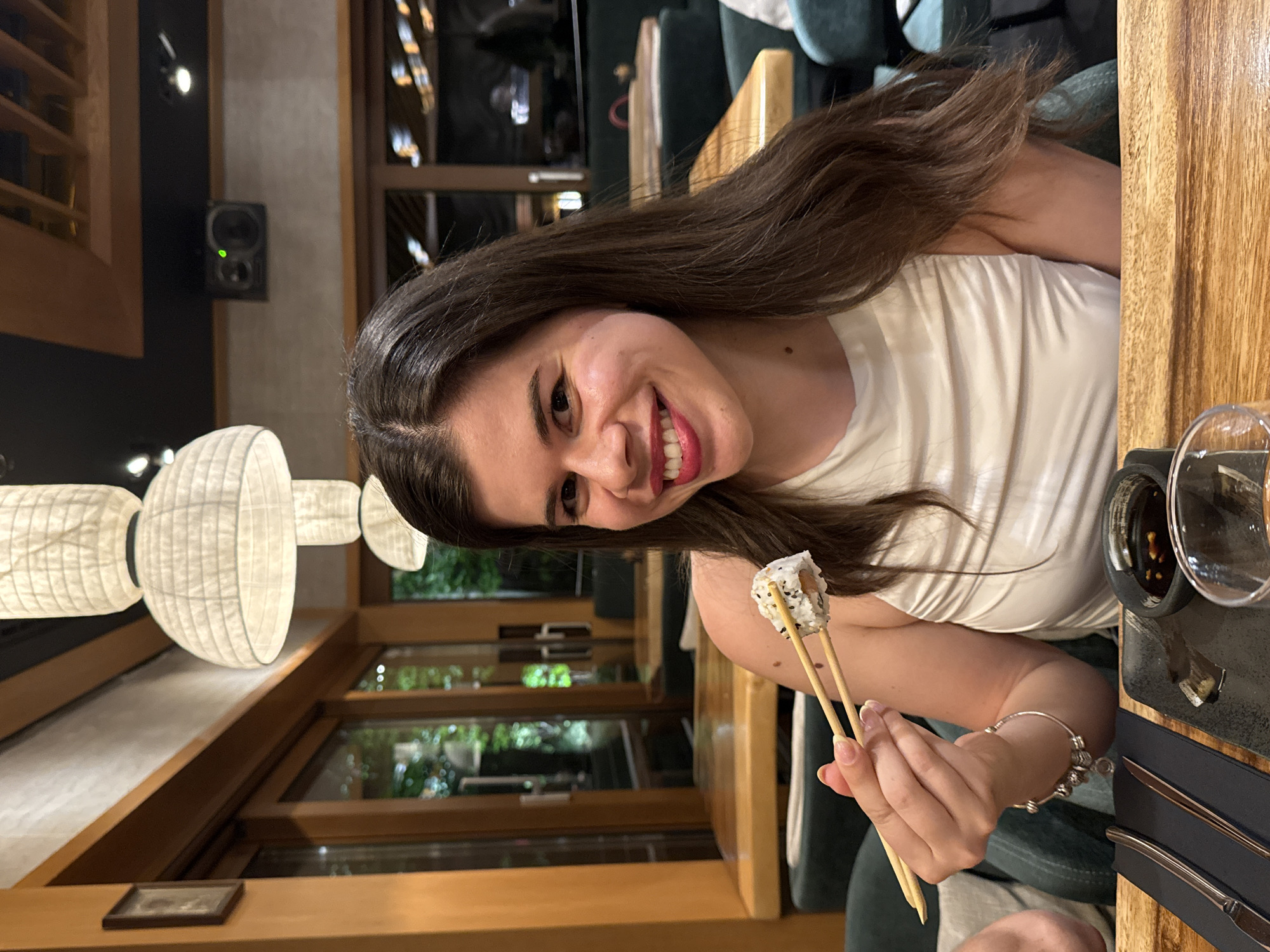 A smiling woman with long brown hair, wearing a white top, holds a piece of sushi with chopsticks at a restaurant. The background suggests an upscale dining establishment, with wooden decor, hanging lights, and large windows. Cutlery and a dish of sauce are visible on the table beside her. While the city of Sofia, Bulgaria, is specified, there is no readily identifiable evidence in the image to confirm this location.