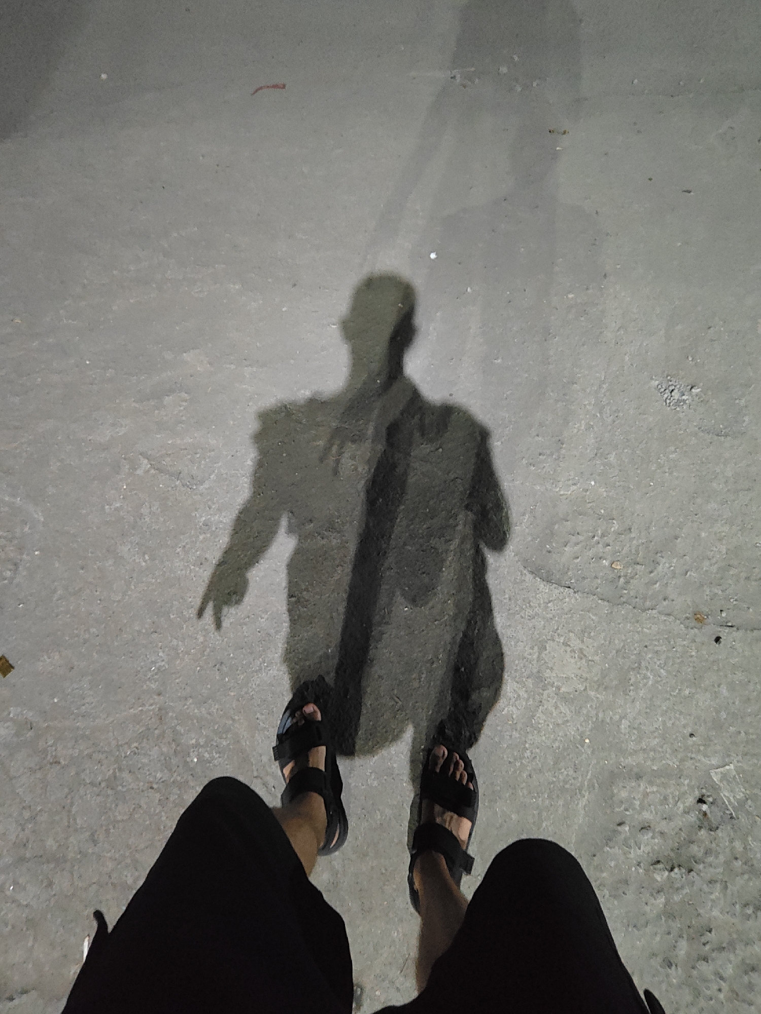 A person stands on a gray, paved surface at night, their shadow cast in front of them. The person is wearing black shorts and black sandals. The shadow is of a person pointing. The location cannot be determined to be Mumbai, India from the image.