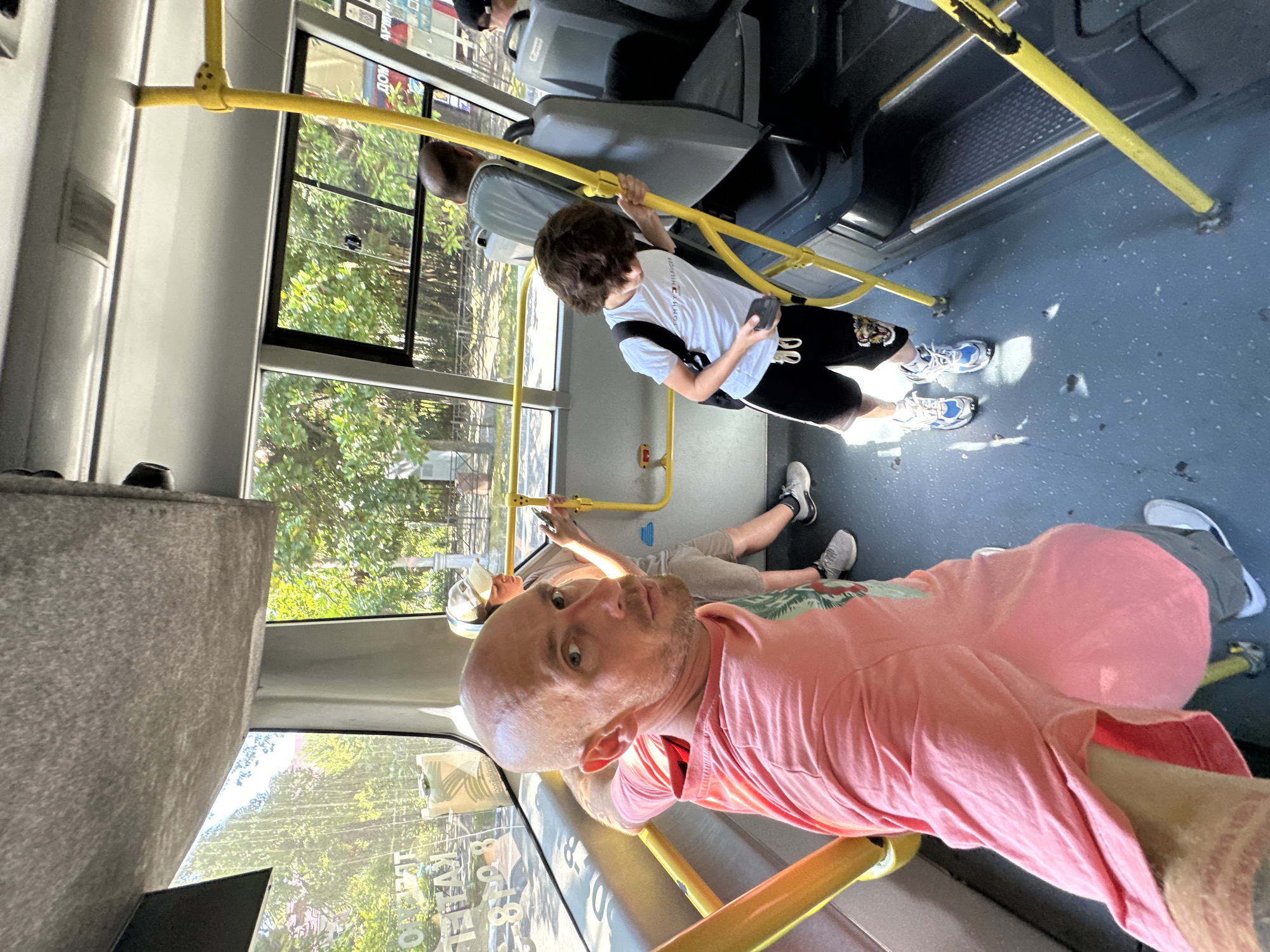 The image captures an interior view of a bus in Sochi, Russia. The bus appears to be on a route with trees visible through the windows, suggesting an urban or suburban setting. A man with a shaved head and a pink t-shirt is in the foreground, looking directly at the camera. He is holding onto a yellow handrail. Behind him, a child with dark curly hair wearing a white t-shirt and black shorts is also holding onto a yellow handrail and looking down at a phone in their hand. Another person, partially visible, is seated further back in the bus. The flooring of the bus is a light blue. The lighting suggests it is daytime.
