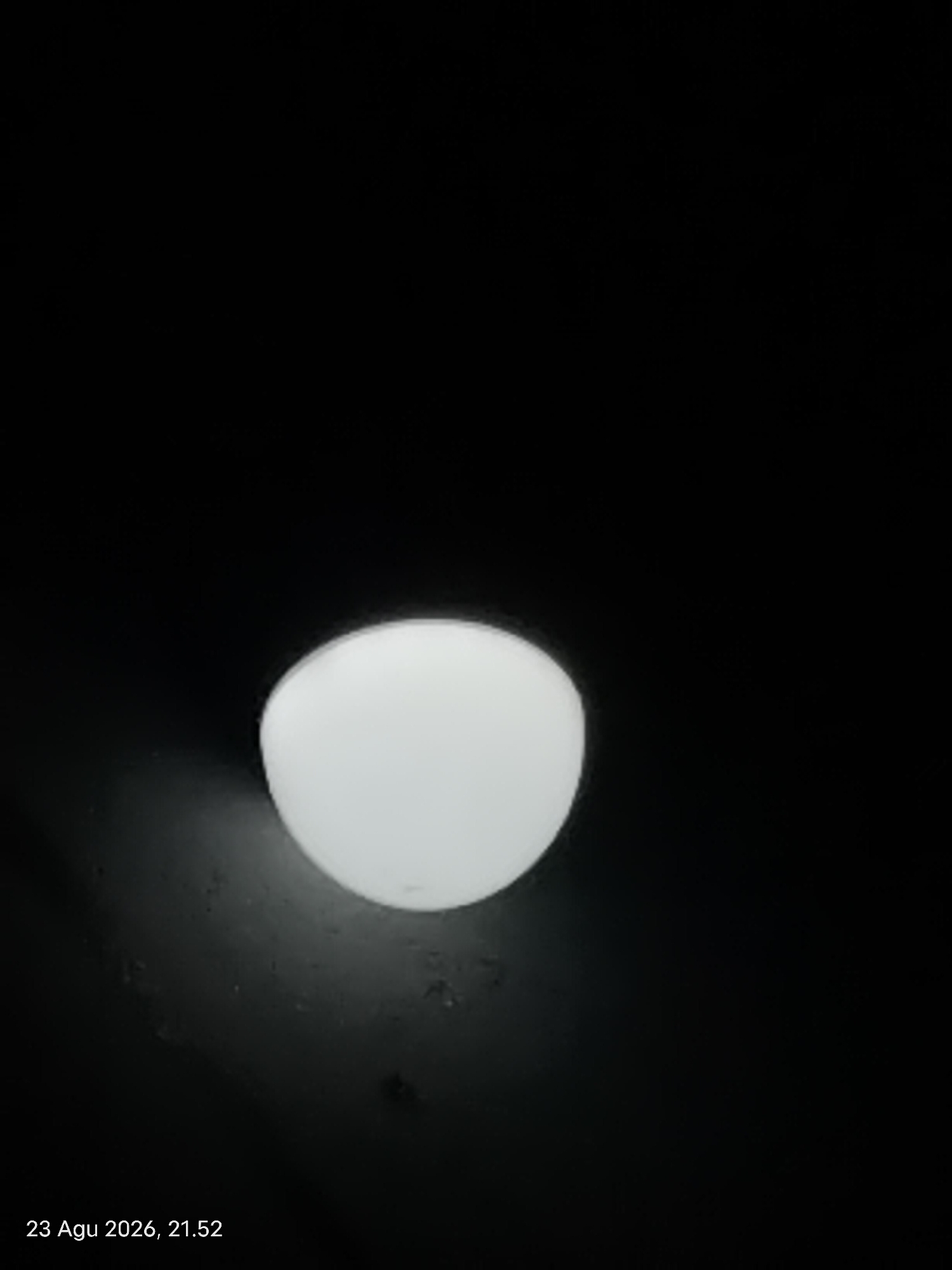A single, circular, white light fixture is illuminated against a black background. The light fixture is positioned in the lower-middle section of the frame. The surrounding area appears to be a dark, textured surface. A timestamp in the bottom-left corner reads "23 Agu 2026, 21.52". The image is dark and lacks any discernible urban or natural landscape features, making it impossible to determine the location within Singkawang, Indonesia. There are no people, actions, or interactions depicted.