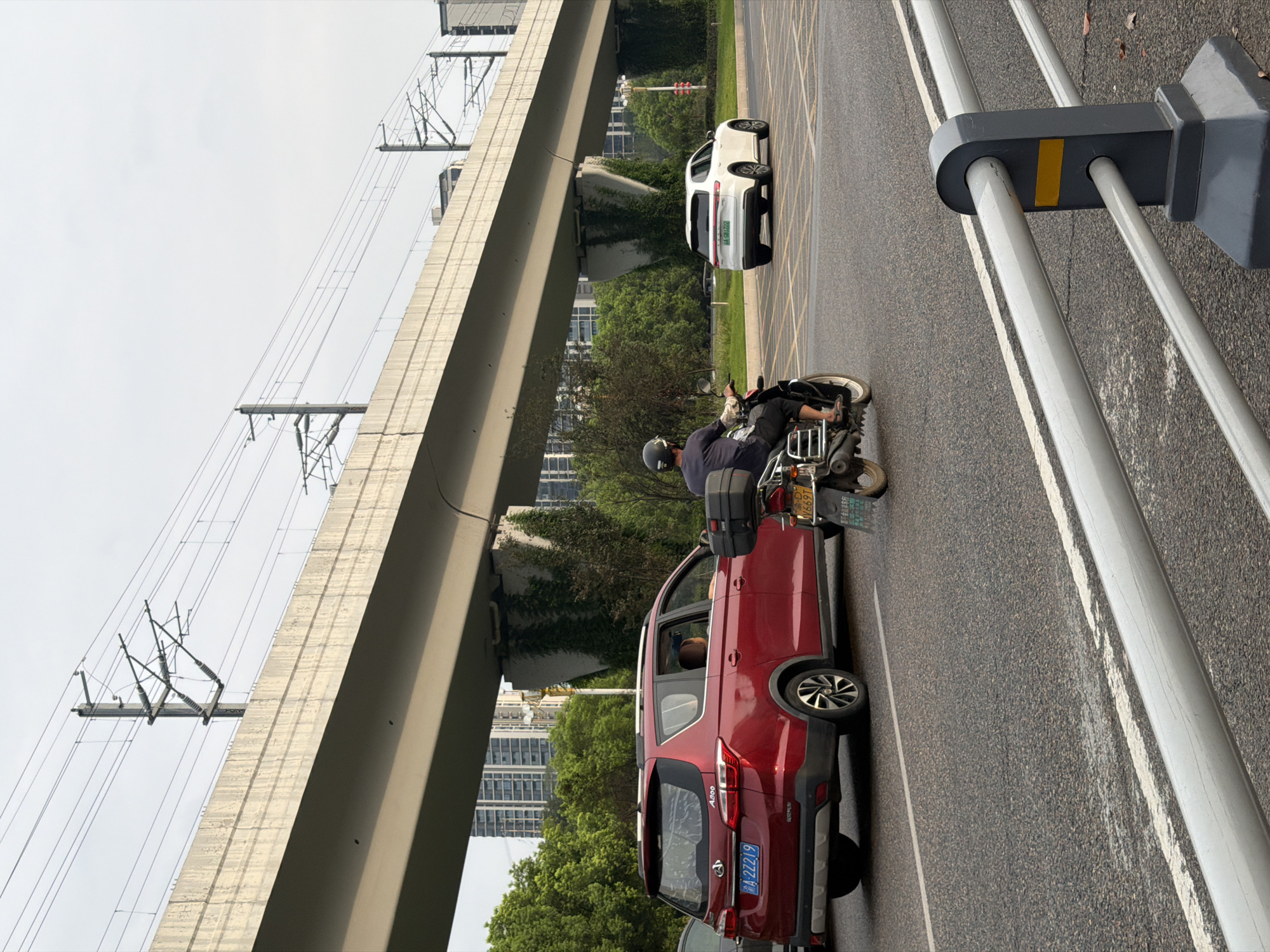 The image depicts a street scene in Jiangjin District, Chongqing, China. An elevated concrete overpass, likely part of a transportation infrastructure like a highway or railway, dominates the upper portion of the frame. Power lines and support structures are visible above the overpass, suggesting an electrified rail line. Beneath the overpass, lush green trees and vegetation line the roadside, with tall residential or commercial buildings visible in the background.
On the road, traffic is present. A red SUV, identified by a license plate ending in "22219", is stopped or moving slowly in the left lane. Inside the SUV, a passenger can be seen. In the right lane, a white SUV, with a license plate ending in "02122", is also present. The main subject in motion is a person riding a motorcycle, heading away from the viewer in the right lane. The motorcycle has a black storage box mounted on the rear and a license plate ending in "6697T". The rider is wearing a dark helmet and casual clothing. The road surface is asphalt, marked with white lines, and a yellow grid pattern indicating a turning lane or intersection area. In the foreground, a metal barrier with gray posts and light-colored horizontal bars is visible, suggesting a pedestrian walkway or divider. The sky is overcast, indicating a diffused light condition.