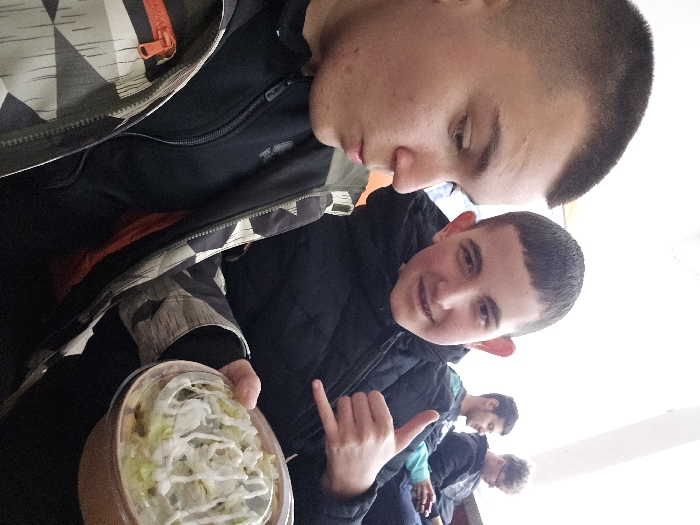 This image portrays four young males seated indoors within an unidentified building in Samokov, Bulgaria, consistent with a casual dining or waiting area. In the foreground, a male with a buzz cut wears a grey, black, and white patterned jacket over a dark zip-up top, looking downward and to his right, with his mouth slightly open. Adjacent to him, a second male with short brown hair and prominent ears, wearing a black zip-up jacket, smiles directly at the camera, displaying dental braces on his upper teeth. He holds a clear plastic container with salad topped with white dressing in his left hand, while his right hand forms a 'shaka' gesture. In the mid-background, a third male with dark hair, wearing a dark jacket with a light blue detail, looks downward and to his right. Further back, a fourth male with light hair and glasses wears a dark hoodie and is also looking downward, likely at a personal device. The immediate surroundings feature plain, bright walls and overhead lighting.