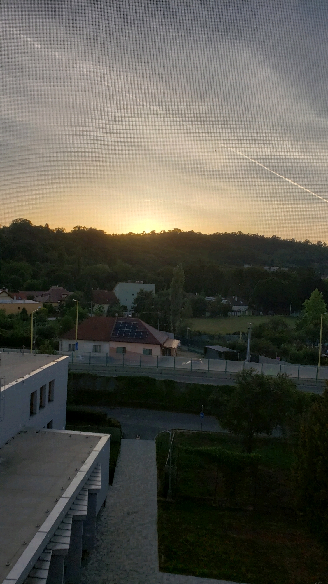 The image captures a view of a suburban landscape at sunset, possibly from a window with a screen. The foreground features the edge of a modern building with a flat, grey roof. Beyond this, a grey paved walkway leads towards a road bordered by a green embankment. Across the road, several houses are visible, one with solar panels on its roof, nestled amongst trees. The background is dominated by a tree-covered hill silhouetted against a pale orange and grey sky, with contrails from aircraft streaking across it. The overall scene suggests a residential area on the outskirts of a city.