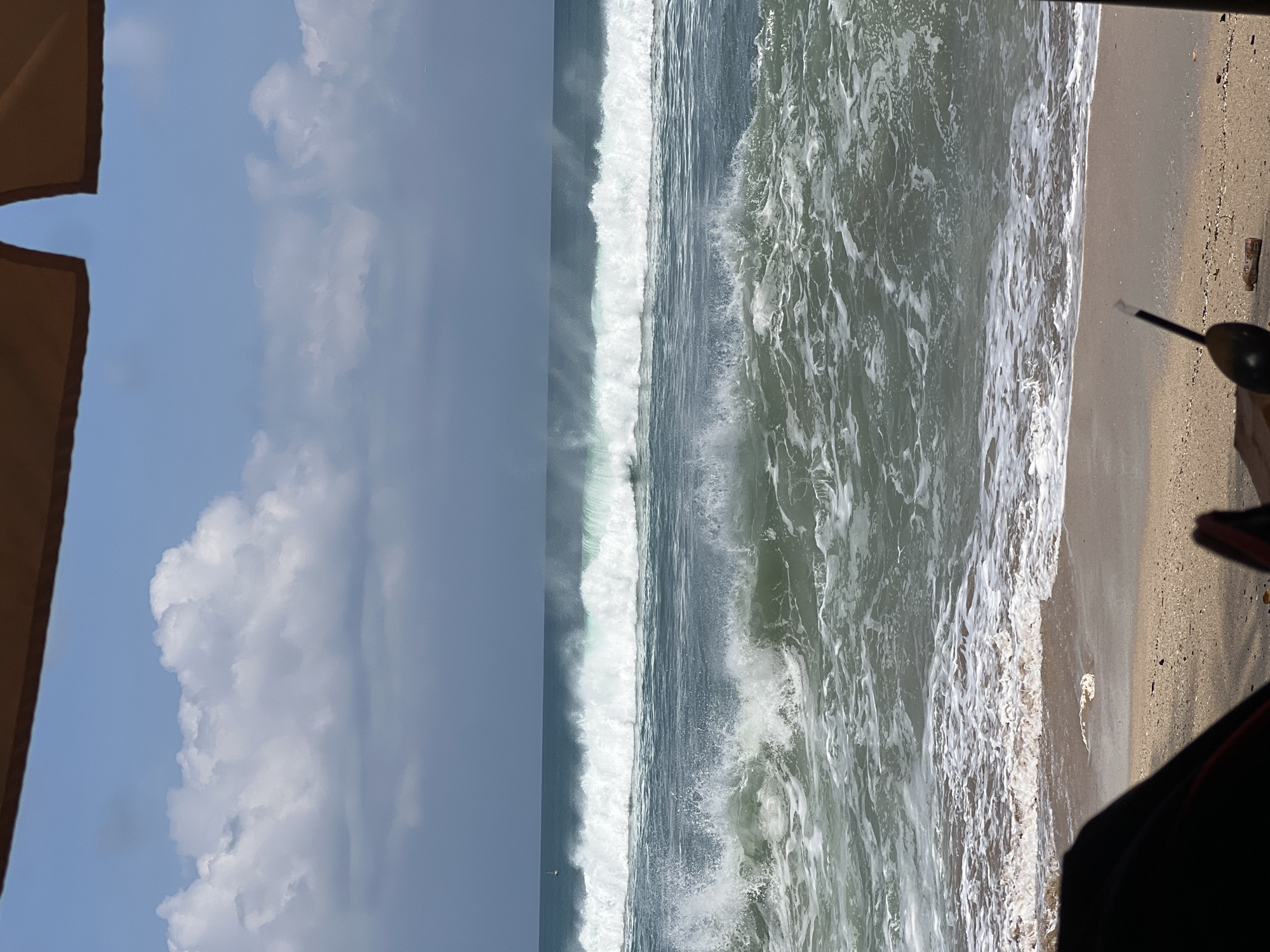 The image shows a view of the ocean from a beach in Kuta, Indonesia. The sky is partly cloudy with large white clouds against a blue sky. The ocean has large waves with white foam. The foreground shows a sandy beach with a part of a beach chair or umbrella visible in the lower left corner. There are some objects on the sand, including what appears to be a bottle. The overall scene depicts a dynamic ocean with significant wave action.