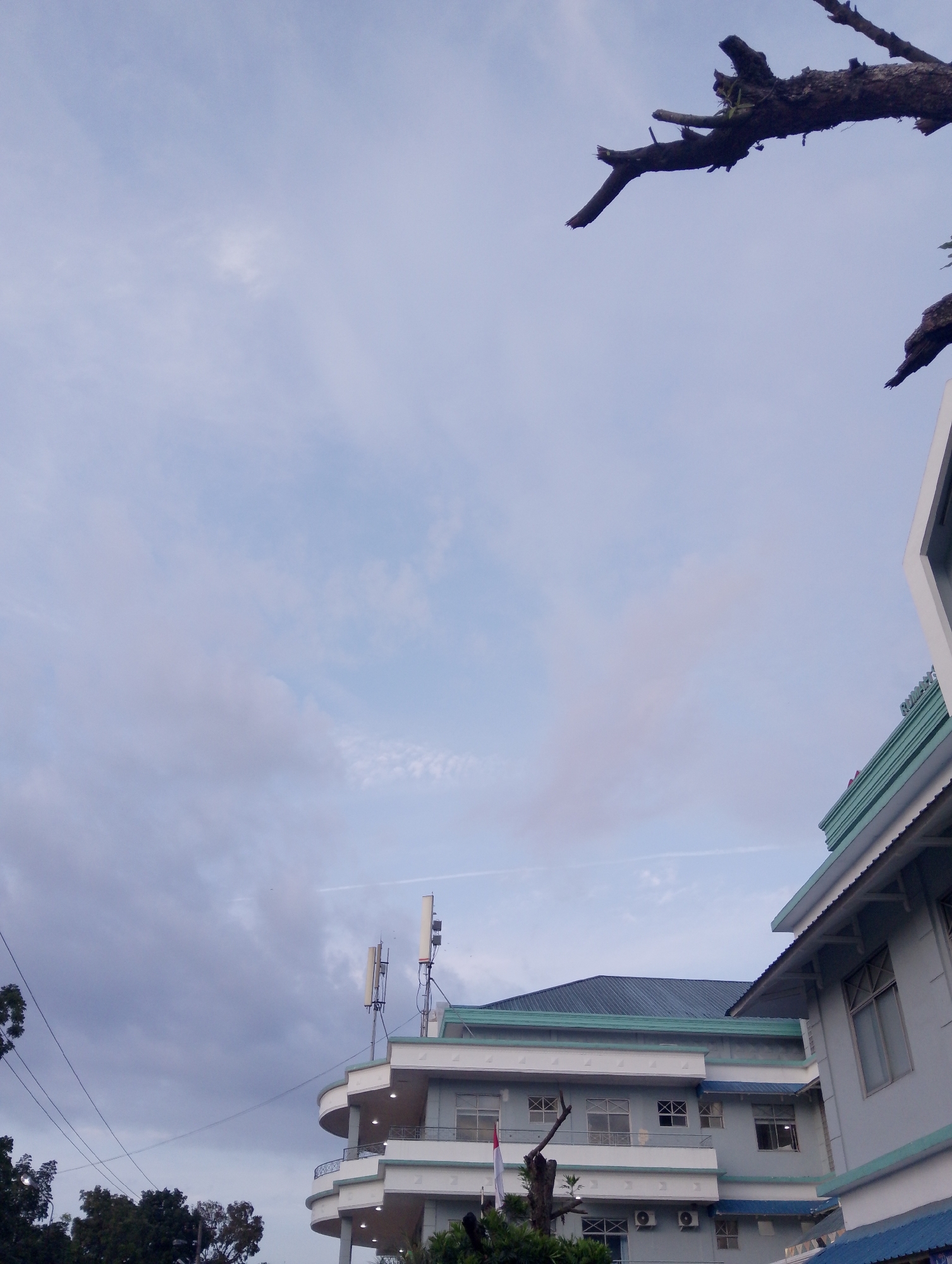 The image shows a building with a light blue facade and a dark blue roof, visible from a low angle. The building has multiple floors with curved balconies and windows. There are two antenna towers on top of the building. Trees and power lines are visible in the foreground. The sky is cloudy and a contrail is visible. The scene appears to be in an urban environment. Based on the visible elements such as the architecture and potential telecommunication infrastructure, it is plausible that this building is located in a city such as Pematang Siantar, Indonesia.