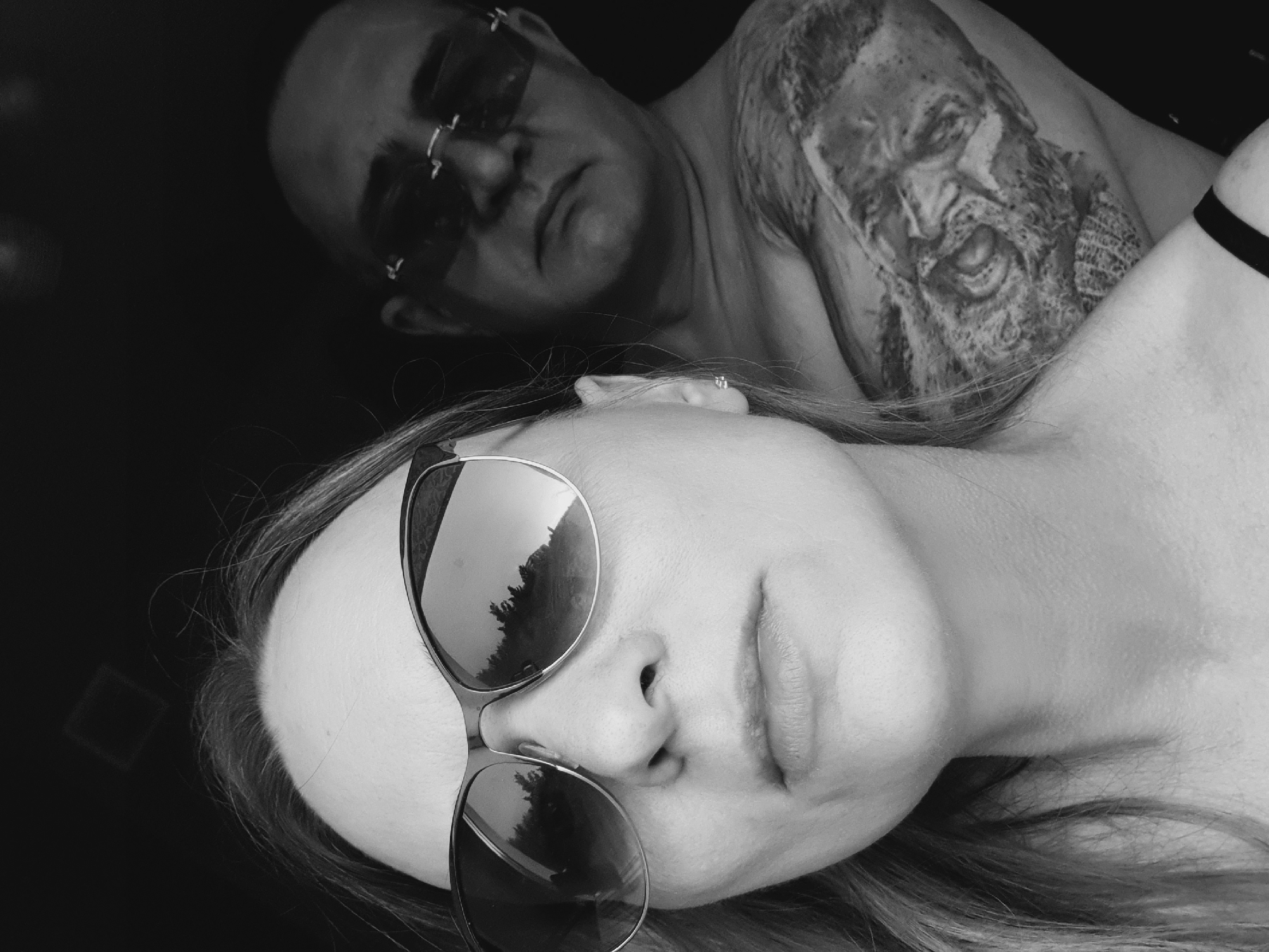 The black and white image depicts two people. In the foreground is a woman lying on her back, facing the camera. She is wearing sunglasses that reflect trees. In the background, a man with sunglasses and a tattoo of a face on his upper arm is looking down at her. There is no clear indication of the location being Ulyanovsk, Russia. The setting appears to be indoors, with a dark background.