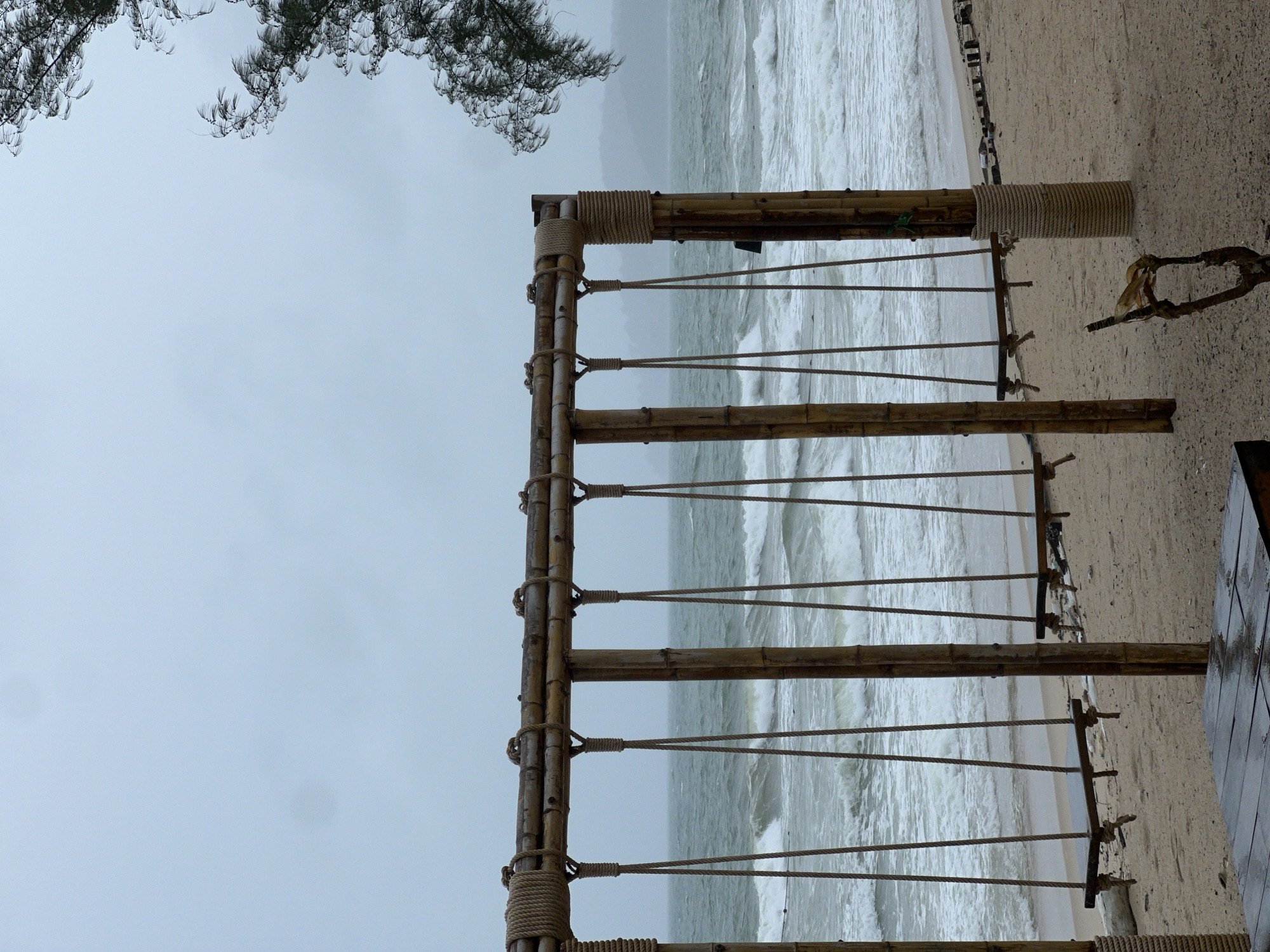 The image displays an outdoor scene at a beach, primarily featuring a rustic bamboo structure in the foreground, with the ocean and an overcast sky behind it. The photograph is oriented vertically, with the beach and sea stretching horizontally across the middle and right side.
A prominent structure made of sturdy bamboo poles is centered in the frame. These poles are lashed together with thick, light-colored ropes, forming a section that resembles a fence, railing, or part of a larger beachfront installation. Several parallel ropes are strung horizontally between the vertical bamboo posts, creating a ladder-like or barrier effect. On the far right, a small portion of a wooden deck or platform is visible, suggesting a man-made viewing area or seating overlooking the sea.
Beyond the bamboo structure, a light-colored sandy beach extends, leading to the ocean. The sea is characterized by numerous white-capped waves, indicating a choppy or rough water condition. The water appears muted in color under the prevailing weather. The sky is uniformly light grey and overcast, suggesting cloudy weather, possibly contributing to the turbulent sea. In the upper left corner, dark, wispy branches of a tree with needle-like leaves, possibly a pine or casuarina, hang into the frame.
The overall mood is tranquil yet wild, underscored by the active ocean and the cloudy sky. No people are visible, and there is no discernible text in the image. The scene captures a natural coastal environment, likely experiencing a period of unsettled weather.