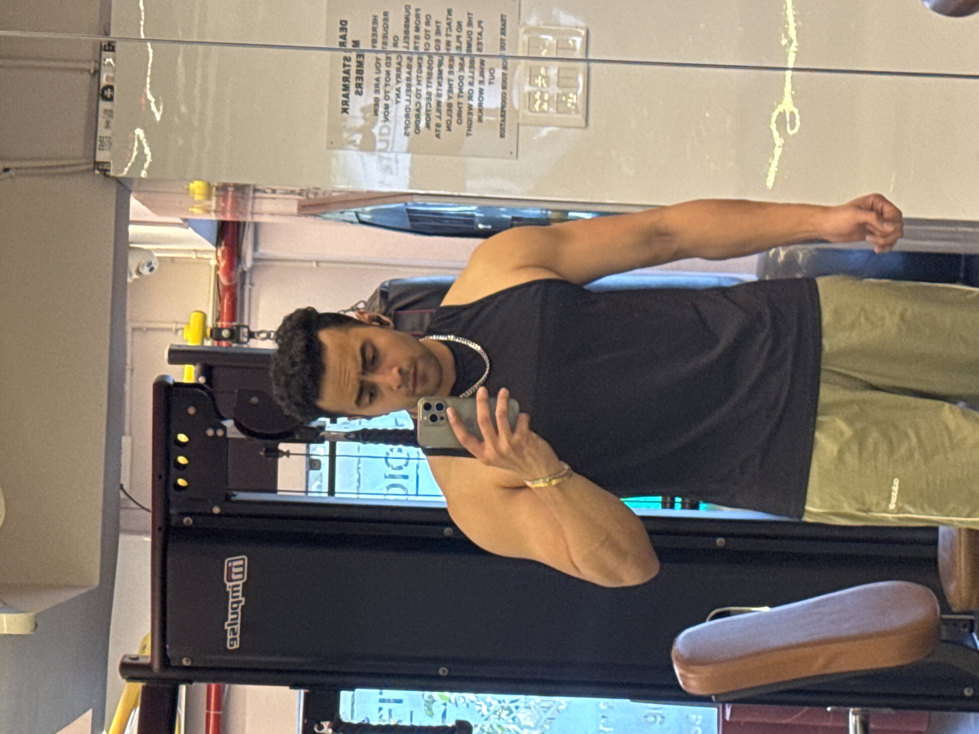 A man is taking a selfie in a gym. He is wearing a black sleeveless shirt and olive green shorts. He has a silver chain necklace and a gold bracelet. He is holding a silver smartphone in his right hand. In the background, there is gym equipment, including a weight machine and a gym seat. There is also a mirrored wall with some writing on it. The writing on the wall appears to be in reverse, as is common in mirror reflections. There is a sign on the weight machine that reads "gaLugm|i". There are also some yellow and red pipes visible in the background, possibly part of the gym's infrastructure. The overall environment suggests a fitness facility. There is no explicit information within the image to confirm the location as Kolkata, India.