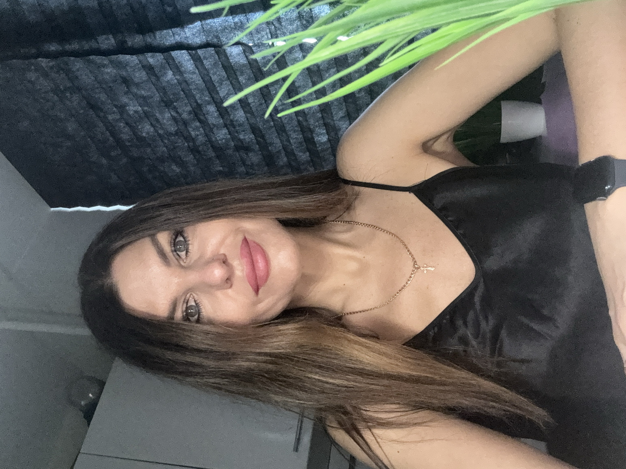 A woman with long, highlighted brown hair is posing indoors. She is wearing a black silk top and a gold necklace with a cross pendant. She has light skin, grey eyes, and pink lips. A black pleated shade is behind her, with an artificial green plant obscuring the top right of the image. The woman appears to be wearing a smart watch on her wrist. The location cannot be determined to be Chaikovsky, Russia, from the given image.