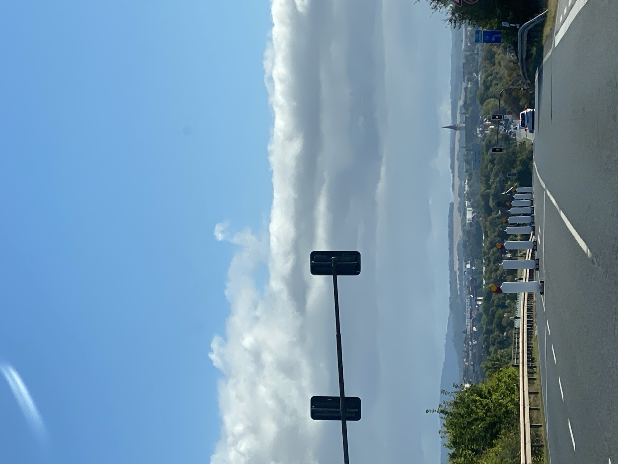 The image depicts a forward view from a vehicle traveling on a road, likely a highway or major thoroughfare, with a town in the distance. The sky is a bright blue with scattered white clouds. In the foreground, the road surface is asphalt with white lane markings. On the left side of the road, a metal guardrail runs along the embankment. Several white traffic cones with yellow tops are positioned on the right side of the road, suggesting construction or a temporary traffic diversion.
Overhead, a traffic light structure spans across the road, with two signal heads visible. Further down the road, a white car is driving away from the camera, and another traffic light can be seen. In the background, a town is visible with buildings and a prominent church spire. The landscape surrounding the town is hilly and covered in trees and vegetation. The overall scene suggests a drive through the outskirts of a European town on a clear day, with indications of roadwork. The presence of the church spire points to the town of Werther, Germany.