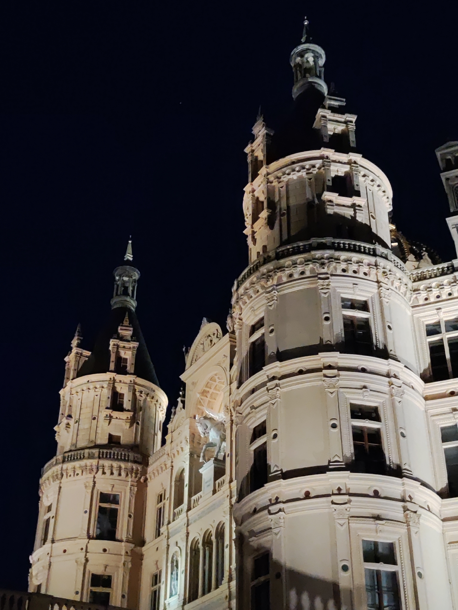 The image shows a low-angle night shot of Schwerin Castle (Schweriner Schloss) in Schwerin, Germany. The castle is illuminated, highlighting its ornate architecture with multiple towers and decorative elements. The sky behind the castle is dark blue, indicating nighttime. The castle appears to be a historic structure with classical design, featuring round towers, arched windows, and detailed stonework. There are no people visible in the image. The scene is static, focusing on the majestic appearance of the castle at night.