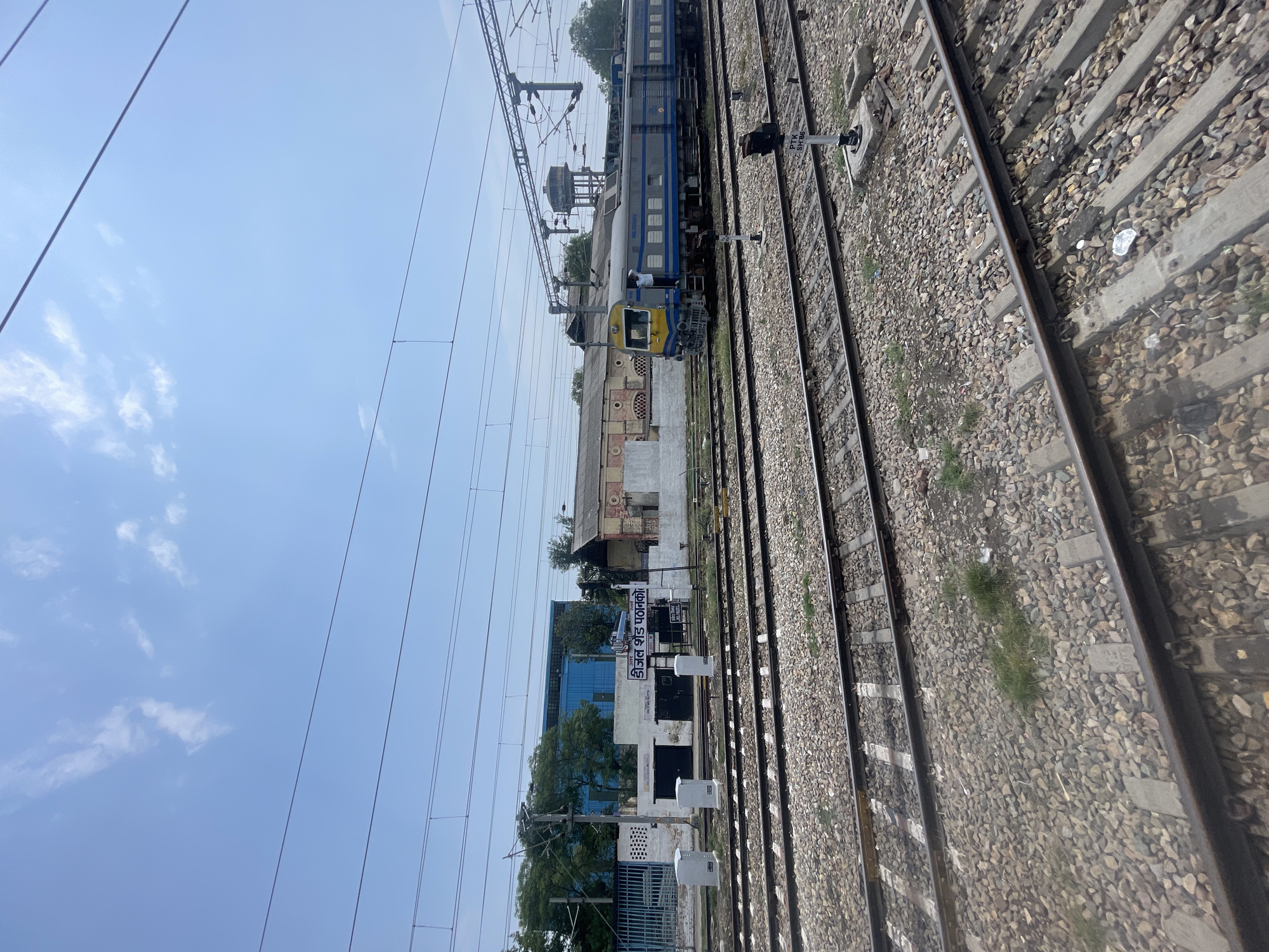The image shows a view of a railway yard in Pathankot, India. In the foreground, multiple sets of train tracks are visible, laid on a bed of gravel and concrete sleepers. The tracks recede into the distance. In the middle ground, a train is parked on one of the tracks. The train is blue and grey with yellow accents on the front engine. A person in a white shirt is standing in the doorway of the engine. Behind the train, there are several buildings. One building is blue and modern, with large windows. Another building is older, with a red brick facade. A sign with Hindi text is visible on the white wall of a low building in the middle ground, identifying the location as "Diesel Shed Pathankot". In the background, overhead electrical wires for the train network are strung across the sky. The sky is blue with scattered white clouds.