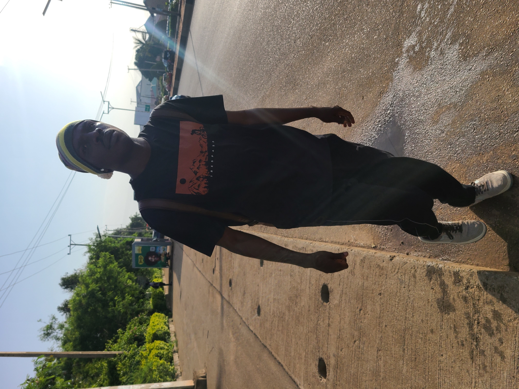 A young Black male walks along a paved sidewalk on the side of a road in Gura Top, Nigeria, under a clear sky with bright sunlight. He is wearing a black t-shirt with an orange graphic depicting mountains and a cyclist, black pants, a multi-colored beanie, and grey athletic shoes. He carries a brown backpack. The sidewalk is made of concrete and has visible circular drainage openings. The road is paved with asphalt and appears wet in places, reflecting the sunlight. Green trees and foliage line the sidewalk. In the background, utility poles and wires are visible, along with some buildings and a few cars on the road.