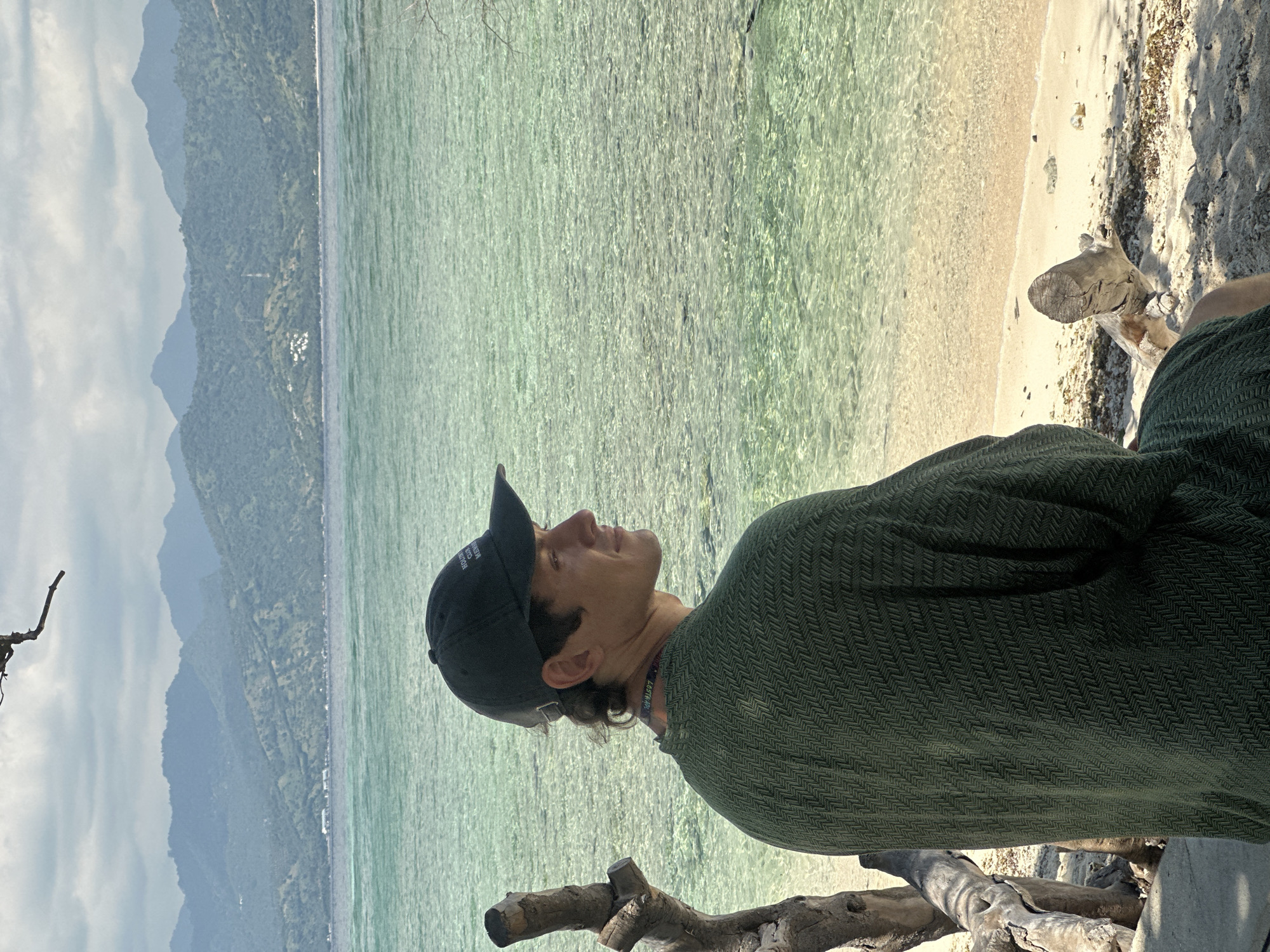 A person wearing a dark baseball cap and an olive-green herringbone shirt is seated on a piece of driftwood near the shoreline. The individual is gazing upwards with their head tilted back. Behind them is a body of water with clear, turquoise-tinted water. A sandy beach is visible along the shoreline. In the distance, a mountainous landscape under a cloudy sky can be seen. The location is in the city of Unknown, Indonesia.