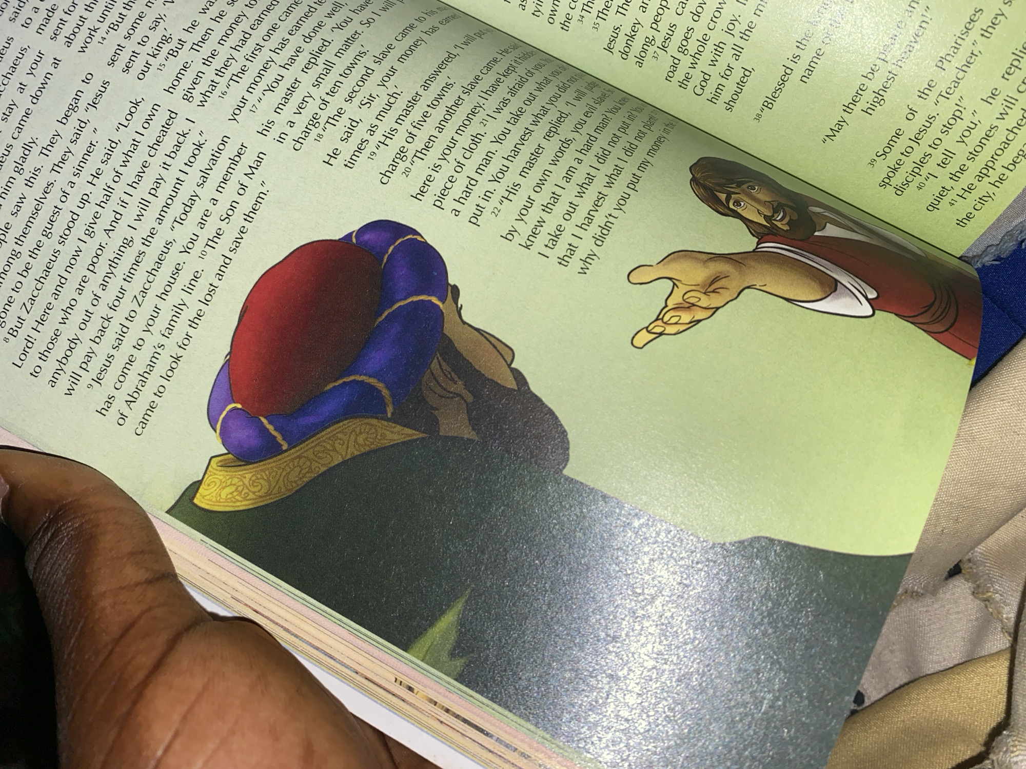 The image shows an open book with text and illustrations. A person's hand is visible in the top left corner, partially obscuring the page. The text is in English and appears to be from a religious scripture, possibly the Bible, with verses numbered and quoted. One illustration depicts a man in a turban and another of a figure identified as Jesus, extending his hand. The pages of the book are yellowed and show signs of age. The image appears to have been taken indoors, with the lighting suggesting a natural light source. There is no information within the image that links it to Jauro Yinu, Nigeria.
