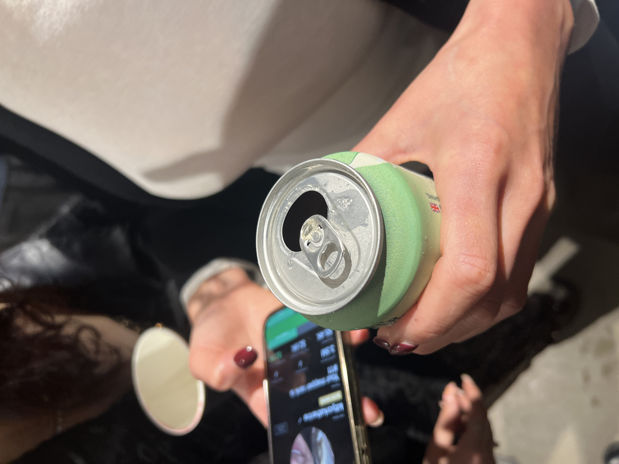 A close-up, high-angle shot captures a person's hands holding a light green beverage can and a smartphone. The right hand firmly grips the can, which is open at the top, showing condensation around the rim. A small flag, resembling the Union Jack, is partially visible on the can's surface, along with some indistinct text that includes "SPRING." The left hand holds a smartphone, displaying an app interface with text, numbers, and a circular profile picture. A circular, possibly reflective object is also held or dangling from the left hand.
The person is wearing a white long-sleeved top or sweater, visible in the upper part of the frame, and dark clothing below. The hands feature dark red nail polish on some fingers. Strong, directional lighting casts noticeable shadows, highlighting the textures of the hands and the can. The background is mostly dark and out of focus, providing no specific location cues. The image appears to capture a casual, candid moment involving drinking and smartphone use.