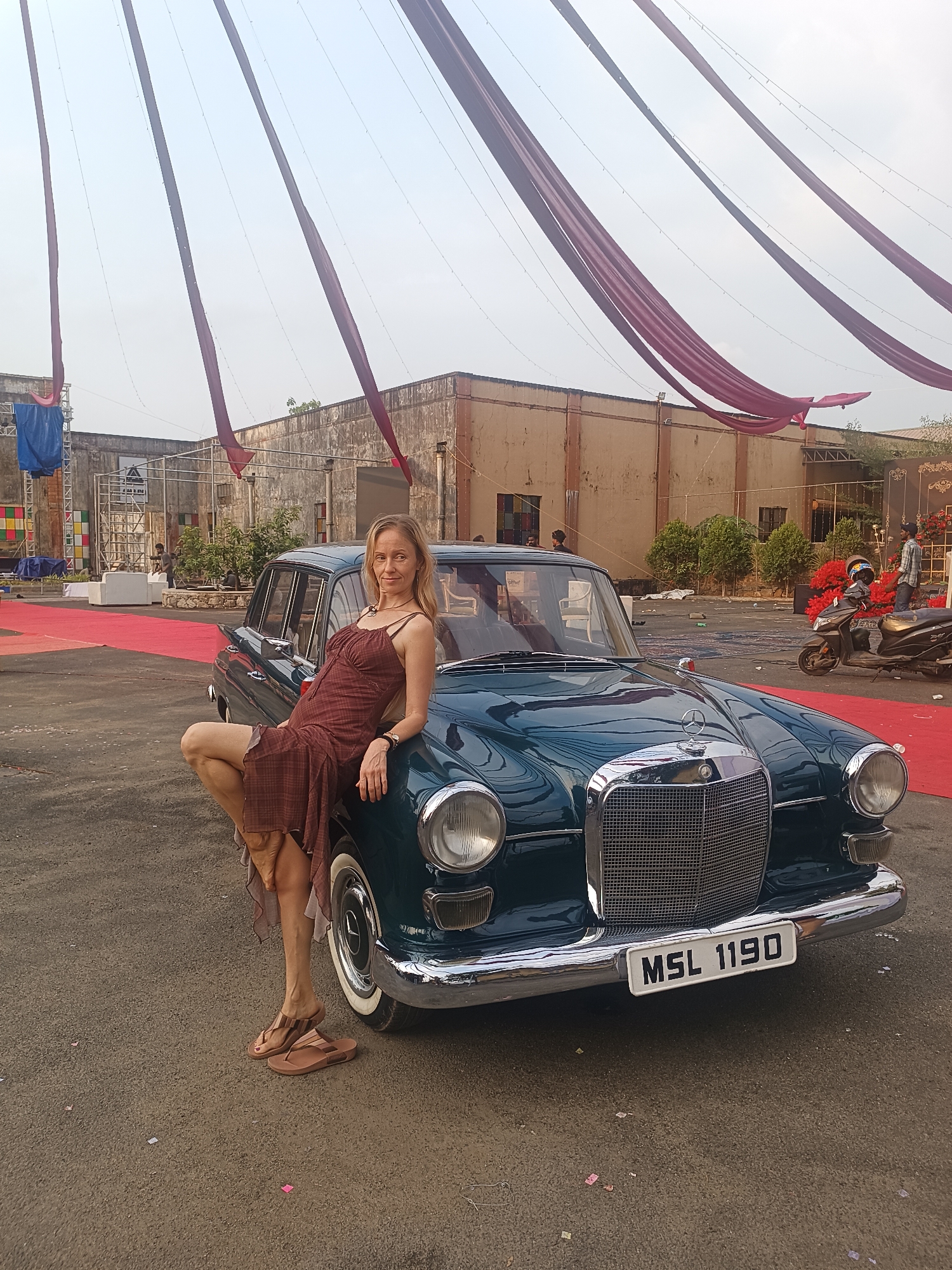 The image captures a fair-skinned woman posing with a vintage dark green Mercedes-Benz sedan in an outdoor setting, likely in Kalamassery, India. The woman, with blonde hair, wears a brown plaid sundress and sandals, leaning casually on the car's hood and front fender. Her left leg is bent, resting on the car.
The scene appears to be an event or photoshoot, characterized by festive decorations. Large maroon fabric drapes stretch across the sky, suspended by thin wires. A red carpet is visible on the ground behind the car, leading towards a complex of concrete buildings with a somewhat industrial or rustic appearance. In the background, various event structures, including a scaffold-like setup draped with blue tarp and decorative elements, are present. Other people are faintly visible in the midground and background, alongside a scooter/moped. The ground is paved, with small pieces of what appears to be paper or confetti scattered.
Notable details include the classic design of the car, possibly a Mercedes-Benz "Fintail" model, with prominent chrome accents and whitewall tires. The car's front license plate clearly displays "MSL 1190." The lighting suggests it is daytime, possibly late afternoon, with a hazy or overcast sky creating soft, diffused light. The overall mood is relaxed and posed.