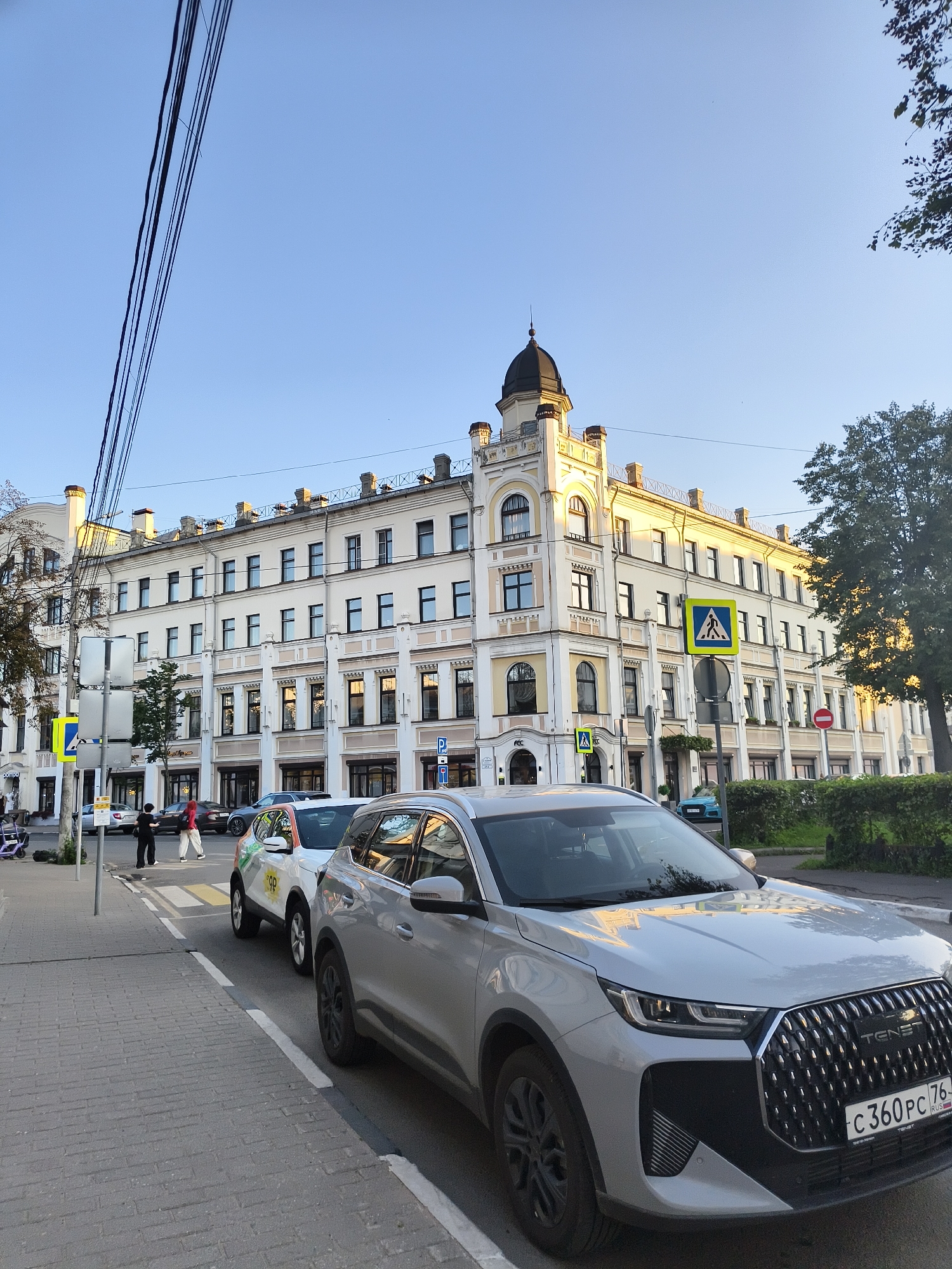 An eye-level, full shot captures a street scene in a city with a building and cars. The building, a pale yellow structure with many windows, dominates the background. It features a dark dome on its roof and architectural details like cornices and decorative elements around the windows. A large tree with green foliage stands to the right of the building. In the foreground, a light silver SUV is parked on the right side of the street. A white car with colorful graphics is parked behind it. Two people are visible on the sidewalk on the left side of the street, one wearing a red top and white pants, the other in dark clothing. The street is paved with asphalt and cobblestones, and there are traffic signs, including a pedestrian crossing sign. Overhead, power lines stretch across the blue sky. The lighting suggests it is late afternoon or early evening, with sunlight casting a warm glow on the building and tree.