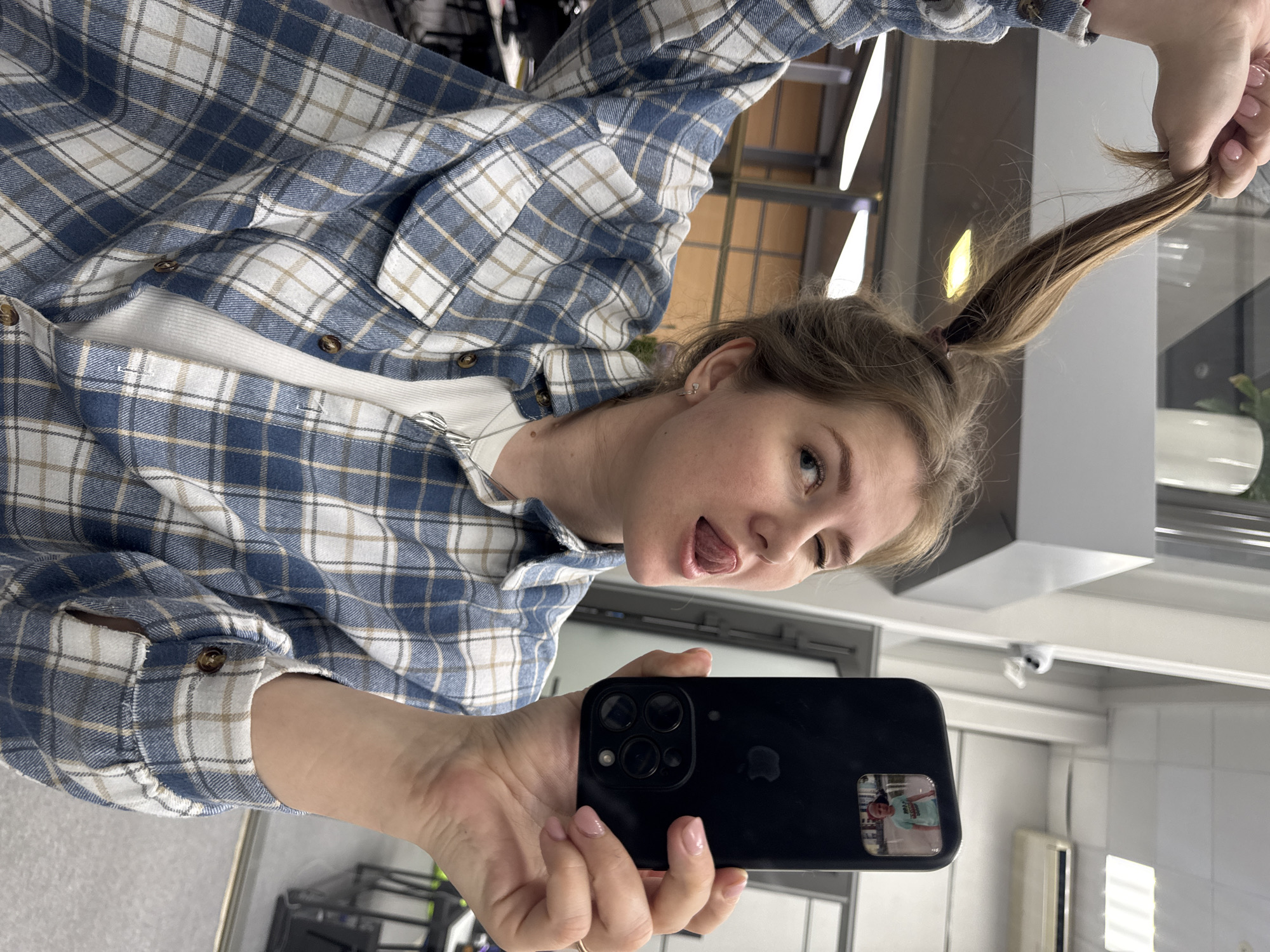 A young woman takes a selfie in a bathroom. She wears a blue and white plaid flannel shirt over a white t-shirt, and her blonde hair is styled in a high ponytail, which she holds with her right hand. Her tongue is sticking out, and she appears playful. She holds a black smartphone, likely an iPhone, in her left hand. A reflection of another person can be seen in the phone's camera lens. The bathroom has white walls and fixtures. Based on the fixtures and appearance of the interior, the location may be a public restroom in Perm, Russia.