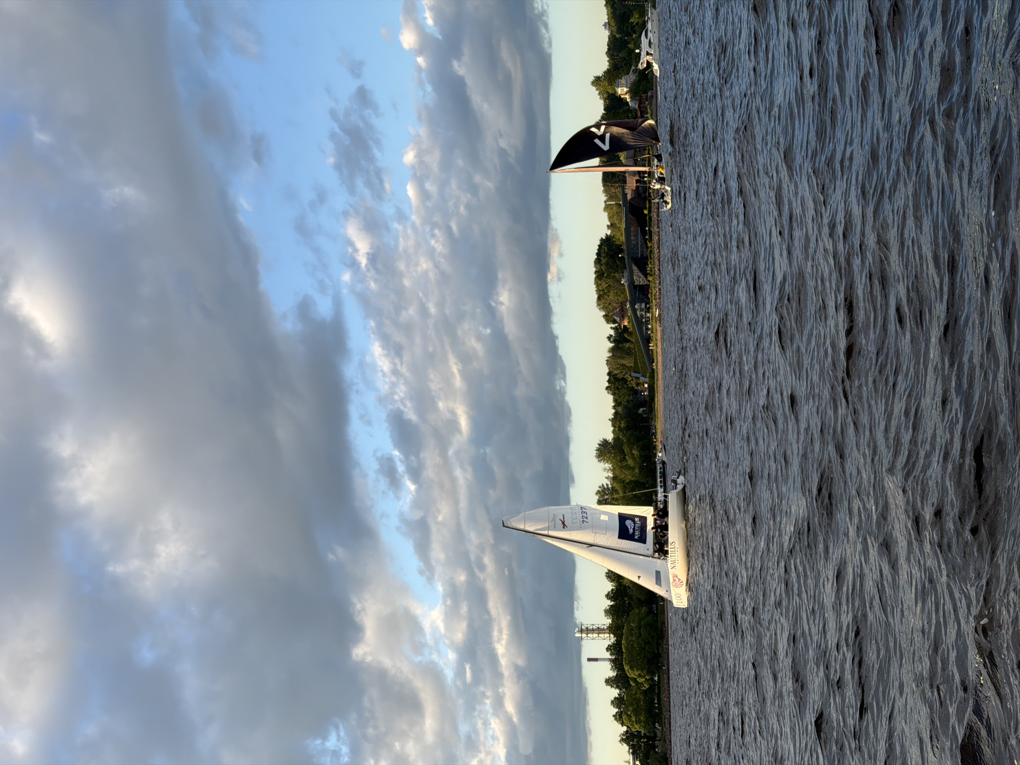 An outdoor, eye-level, full shot captures a body of water with multiple sailboats and a distant cityscape under a cloudy sky. The water is dark and choppy, with visible ripples and waves. In the foreground, a white sailboat with a tall mast and a partially unfurled white sail is positioned on the right. The sail has red and black lettering that includes "100" and "NAUTILUS SAILING". Several people are visible on the deck of this sailboat.
To the right of the white sailboat, a second sailboat with a large black sail patterned with a white chevron design is sailing. This boat also has people on deck. Further to the right, a white motor yacht is visible.
In the background, a tree-lined shore is visible, with a few buildings scattered among the trees. A distinctive industrial tower with red and white markings and a smaller tower with antennas are also visible on the left side of the frame, suggesting an industrial area within or near the city. The sky above is filled with dramatic, textured clouds, with patches of blue sky visible between them, indicating either sunrise or sunset lighting. The overall impression is a scenic view of sailing activity on a large body of water with the urban landscape of Saint Petersburg in the distance.