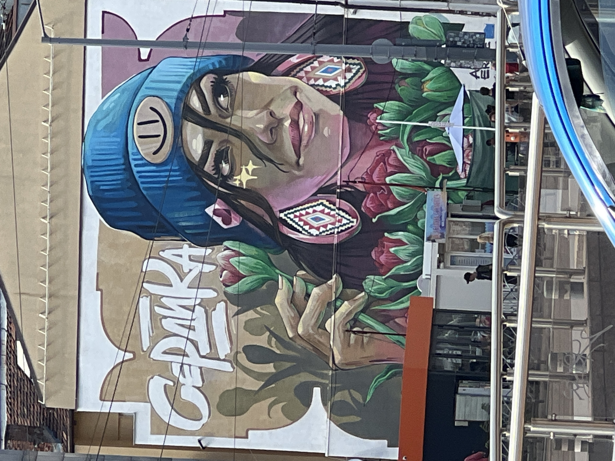 The image shows a large mural on the side of a building in Sofia, Bulgaria, depicting a young woman wearing a blue beanie with a smiley face emblem. She has dark hair, striking eyes with dramatic makeup, and large, patterned earrings. She is holding a bouquet of pink and red flowers. The mural also features the word "SERDIKA" in large white lettering, an ancient name for Sofia. In the foreground, there are people walking and standing around a street-level structure with glass windows, possibly a kiosk or shop. Metal railings are visible, separating the sidewalk from a lower level. A blue car is partially visible in the bottom right corner. The building itself has a tiled roof and appears to be multi-story. Electrical wires and a utility pole are present near the mural. The overall scene depicts a vibrant urban environment with street art.