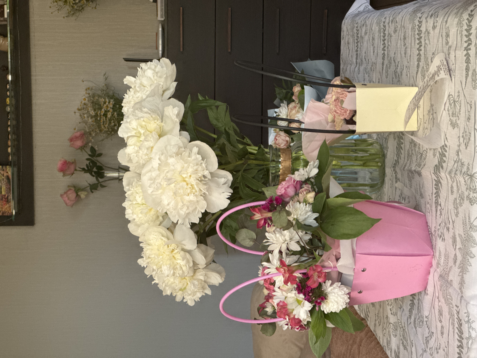 The image shows a still life arrangement of flowers on a surface with a floral pattern. There are three bouquets of flowers. One bouquet features large white peonies in a clear glass vase. Another bouquet is arranged in a pink bag, and a third bouquet is arranged in a yellow bag. The pink and yellow bags are each topped with handles. The background includes a gray wall on the left and a dark brown cabinet in the center. A framed picture is partially visible on the far left of the image. The location is Unknown, Unknown.
