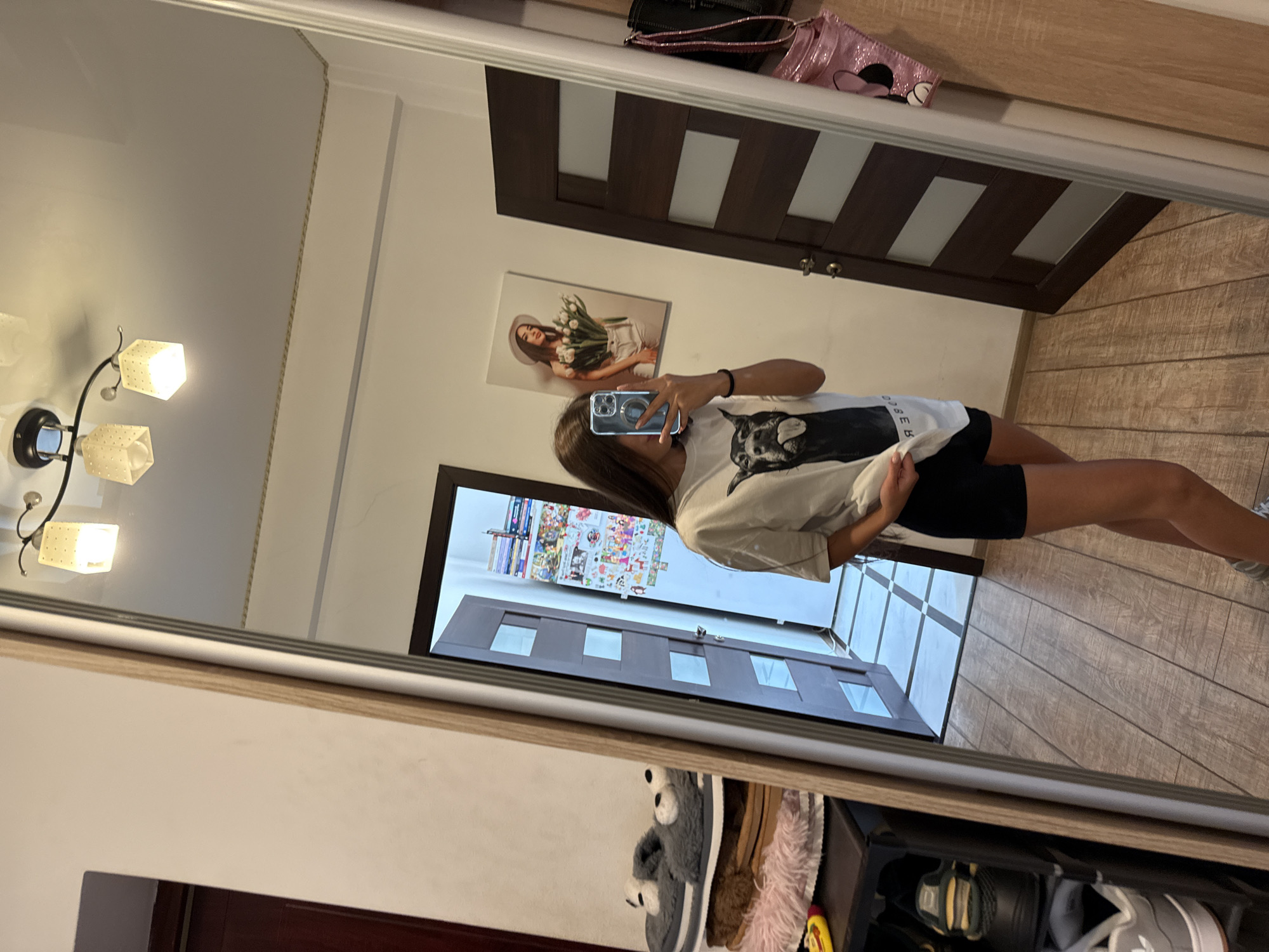 A young woman is taking a selfie in a mirror. She is wearing a white t-shirt with a dog print and black shorts. In the mirror, a wall is visible with a picture of a woman holding tulips, a dark brown door, and a rack of shoes. The floor is tiled. The overall setting appears to be a domestic interior.