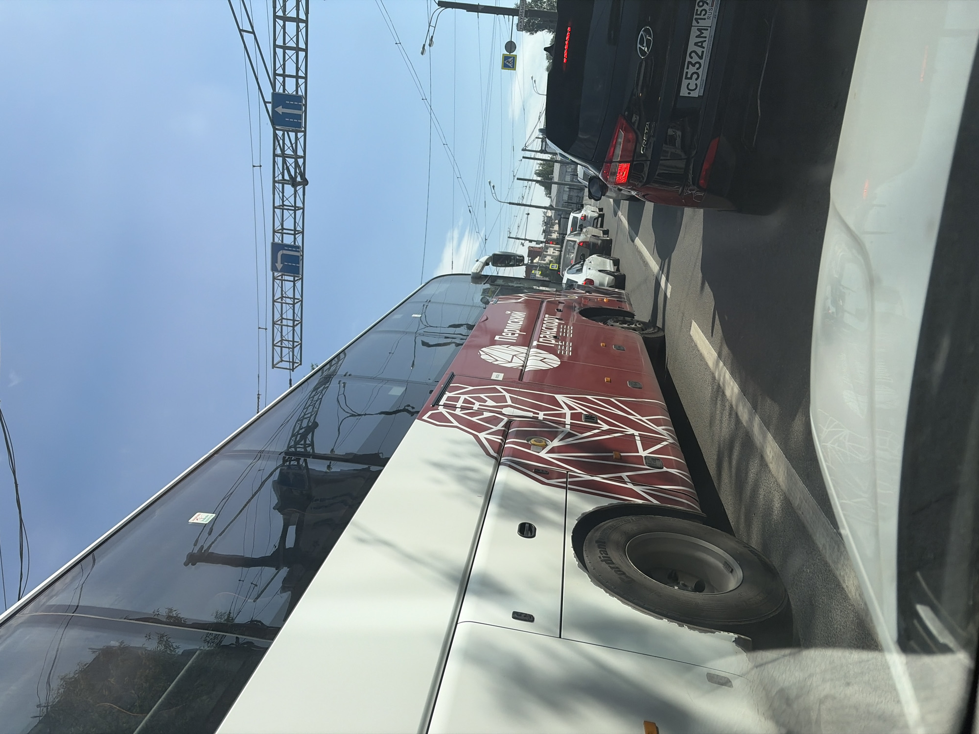 The image shows a view from inside a car in Perm, Russia. The camera is pointed out the passenger side window. The main subject is a large passenger bus with the words "Пермский Транспорт" ("Permian Transport") painted on its side in Russian. The bus has a maroon and white geometric design. To the right of the bus is a black car with the license plate C532AM 159 (Russian plates). Other vehicles are visible further ahead. Overhead, there are wires and utility poles with signs. The sky is blue and sunny. The image captures a typical urban traffic scene.
