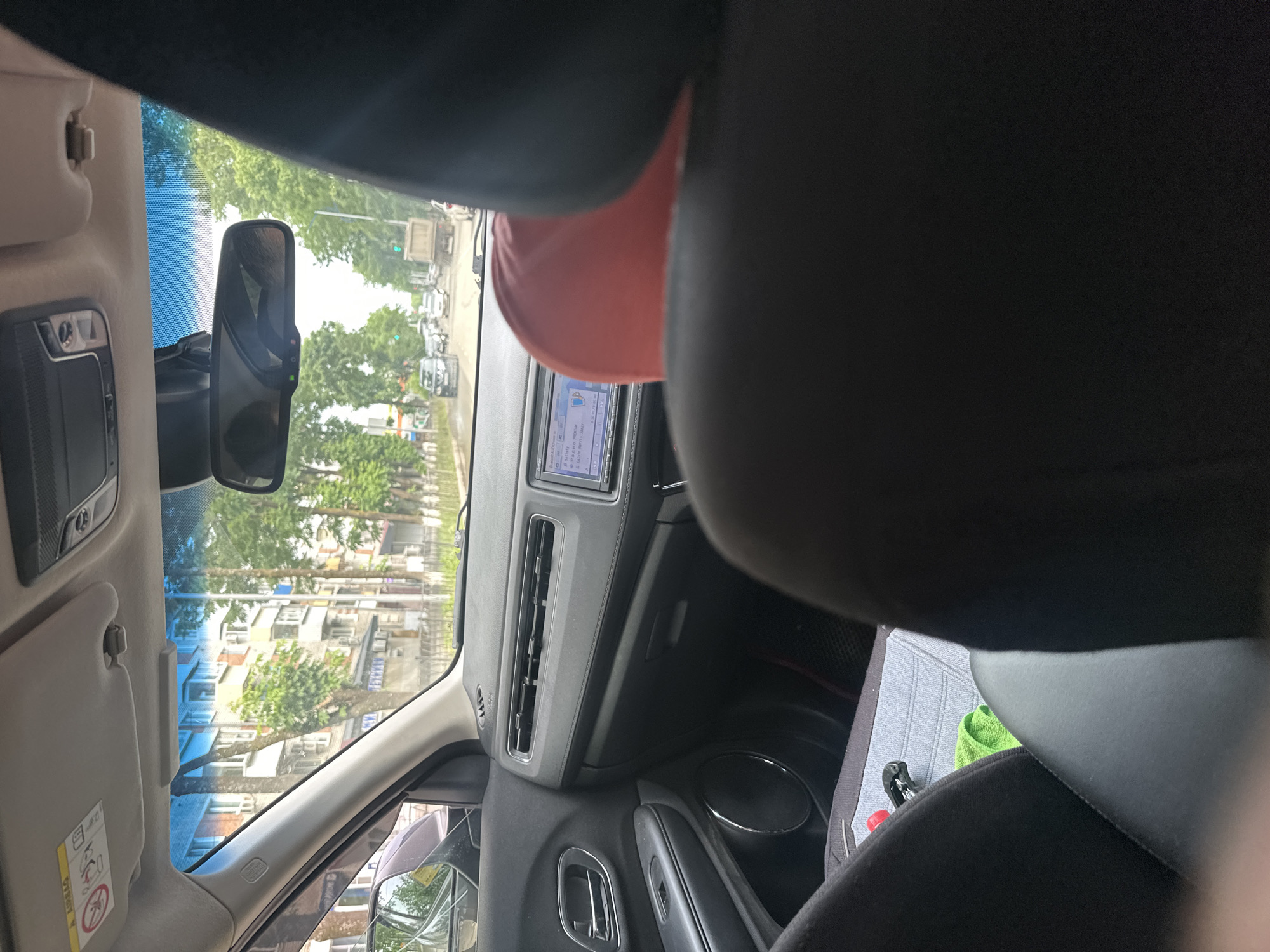 The image depicts the interior of a car, likely a Honda HR-V, as viewed from the rear passenger seat, looking forward. The car is in motion on a street in Birobidzhan, Russia, indicated by the architecture and vehicle models. The dashboard features a multimedia system displaying song titles and artist names, suggesting the car's audio system is active. A person wearing a red shirt is seated in the front passenger seat, partially obscuring the view of the dashboard. The exterior view through the windshield shows a moderately busy street with trees, buildings, and other vehicles, including a dump truck. The overall lighting suggests it is daytime.