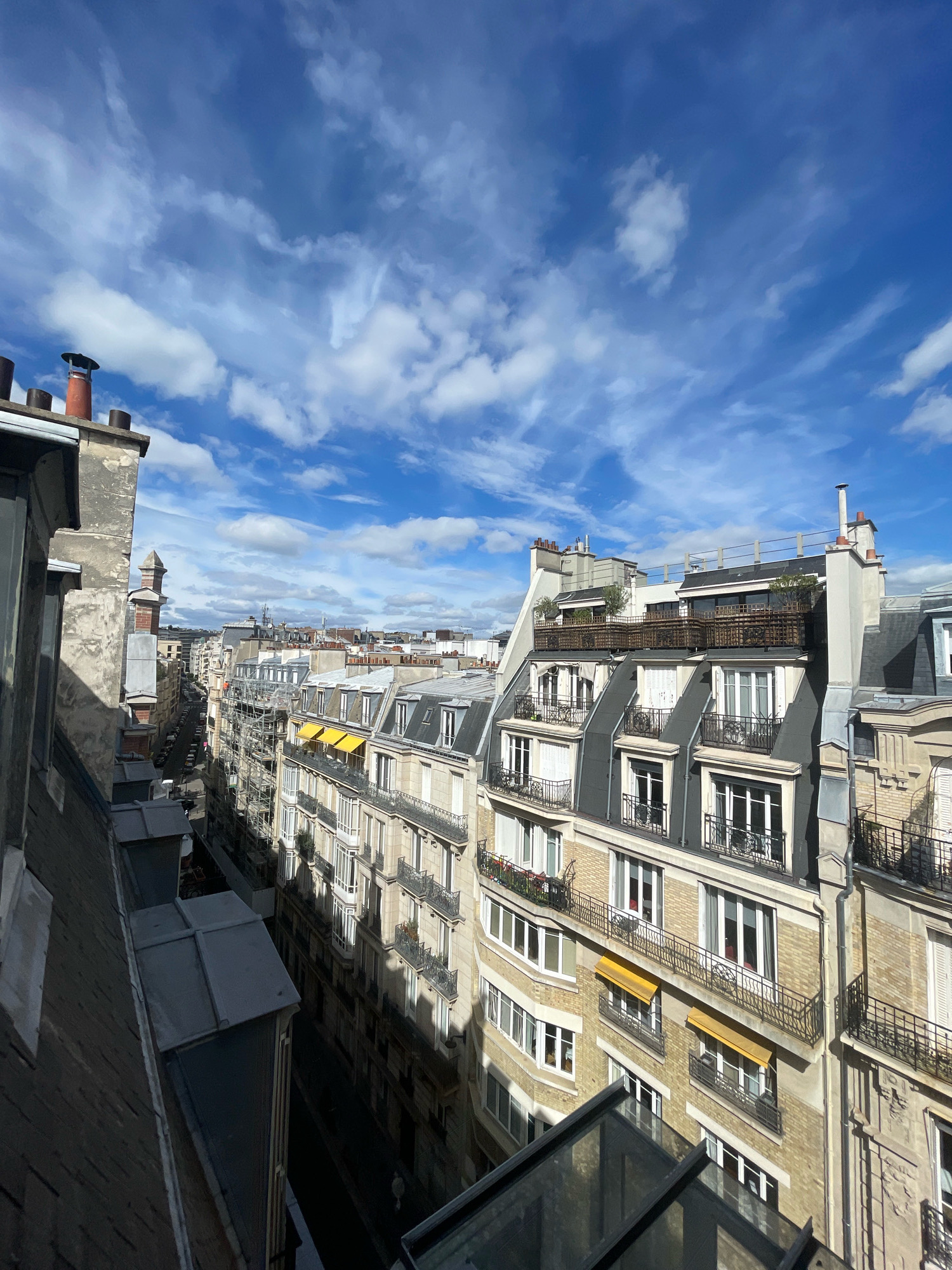 The image captures a view of Parisian rooftops and a street below on a sunny day with a partly cloudy sky. In the foreground, a dark, sloped roof with metallic sheeting dominates the left side of the frame, extending into the bottom half. To the right of this roof, a tall, grey building with visible window frames and a dark chimney rises.
Beyond the immediate foreground, a street lined with Haussmann-style apartment buildings stretches into the distance. These buildings are characterized by their cream-colored stone facades, mansard roofs with dormer windows, and wrought-iron balconies. Several buildings feature retractable yellow awnings over some windows. Scaffolding is visible on at least one building, indicating ongoing construction or renovation.
The street below appears relatively narrow and is lined with parked cars. There are a few indistinct figures walking on the sidewalk. The sky above is a vibrant blue with scattered, wispy white clouds. The overall scene suggests an urban environment in Paris, likely a residential area.