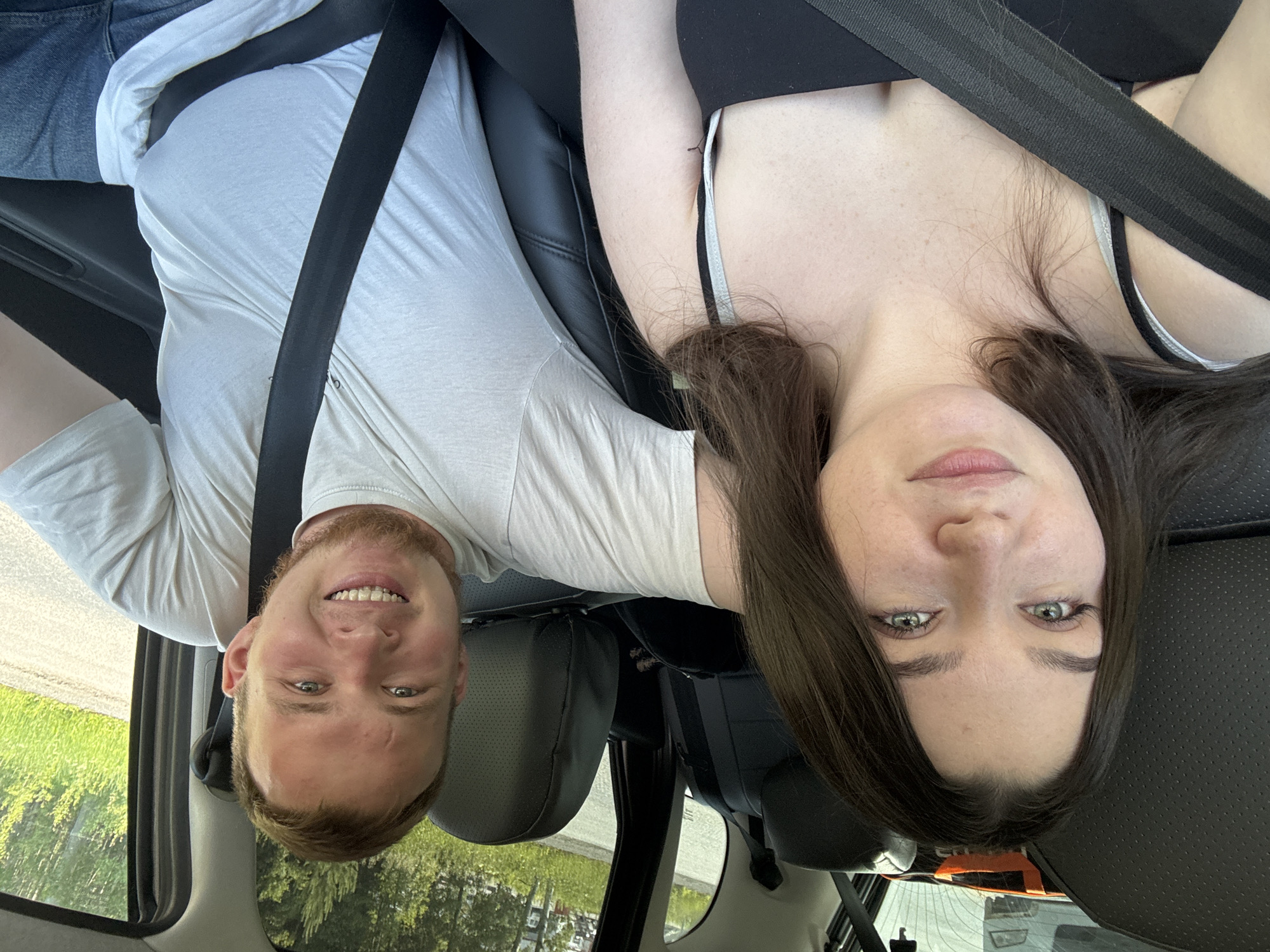 In Pesterevo, Russia, a young couple is seen inside a car, likely a sedan, with leather-like seats. The woman, with long dark hair and light eyes, is in the foreground on the left, looking towards the camera with a slight smile. She is wearing a black top and a seatbelt. To her right, the man, with short blonde hair and a beard, is also looking at the camera, smiling broadly. He is wearing a light-colored t-shirt and a seatbelt. The car's interior is visible, with the dark seats and the orange object on the back seat. Through the car windows, a glimpse of the Pesterevo environment is visible, with trees and greenery indicating an outdoor setting, possibly a road surrounded by nature. The overall impression is of a candid photograph taken during a car journey.