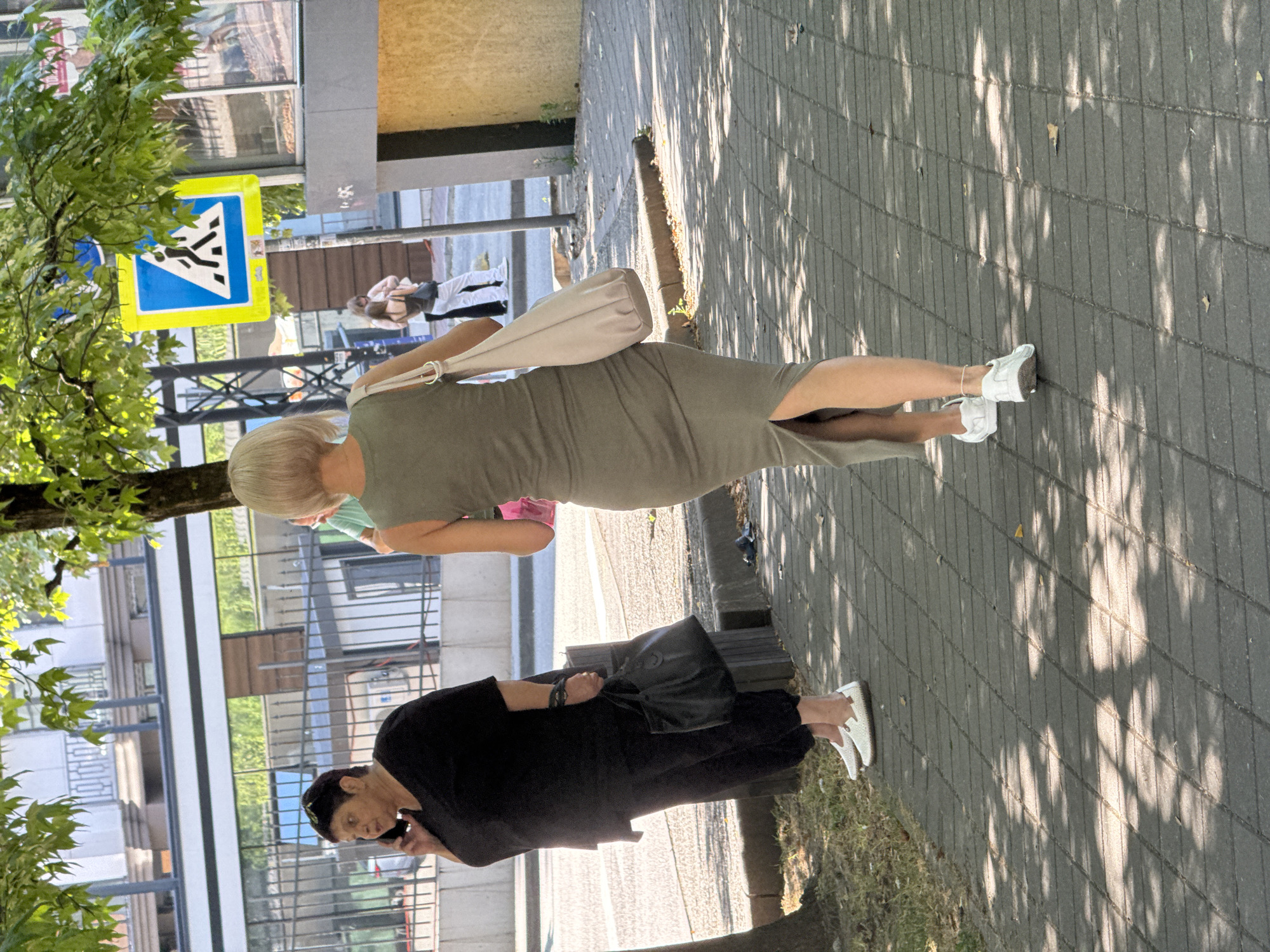 A medium shot shows two women on a paved sidewalk in Sochi, Russia, bathed in dappled sunlight. The woman on the left, dressed in a black top and pants, is on her phone, holding it to her left ear with her right hand and carrying a black handbag in her left. She is wearing white slip-on shoes. To her right, a woman in a fitted olive green midi dress and white sneakers walks away from the camera. She carries a beige tote bag on her right shoulder. The background includes a tree, a pedestrian crossing sign, and buildings with glass facades. The ground is paved with grey rectangular stones.
