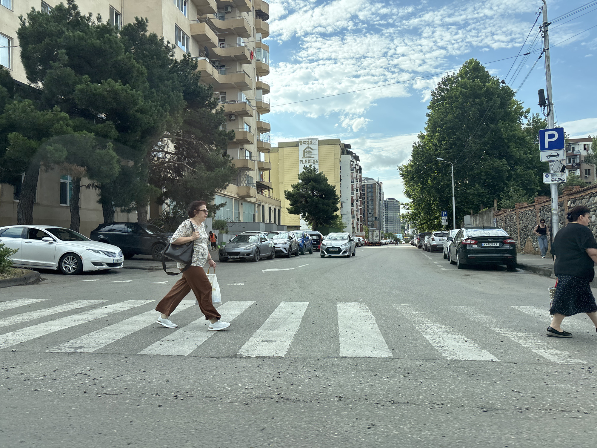 The image depicts a street scene in Tbilisi, Georgia, specifically in the Vake district. In the foreground, a woman with glasses and brown pants walks across a pedestrian crossing, carrying shopping bags. Several cars are parked along the street and also drive in the distance. The background shows multi-story buildings with balconies, as well as trees lining the street. A parking sign is visible on a utility pole on the right side of the image. The sky is partly cloudy.