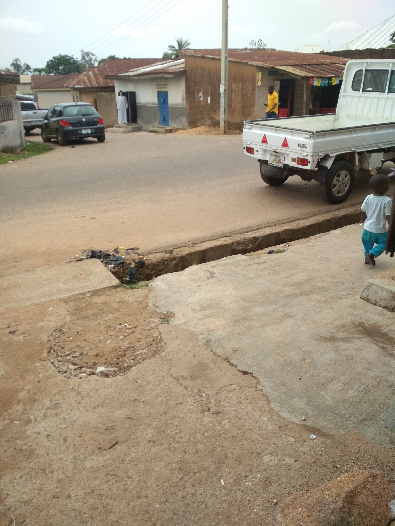 The image shows a street in Unknown, Unknown. A white truck is parked on the right side of the road. A young child is walking on the sidewalk next to the truck. A man in a yellow shirt is standing in front of a building across the street. Further down the road, a person in a white robe is standing in front of a blue door. A black car and a white pickup truck are parked behind them. There is a ditch filled with trash next to the road and in front of the sidewalk. The road and sidewalks are damaged. Buildings are constructed from concrete and covered with roofs of corrugated metal. Power lines are visible in the sky.